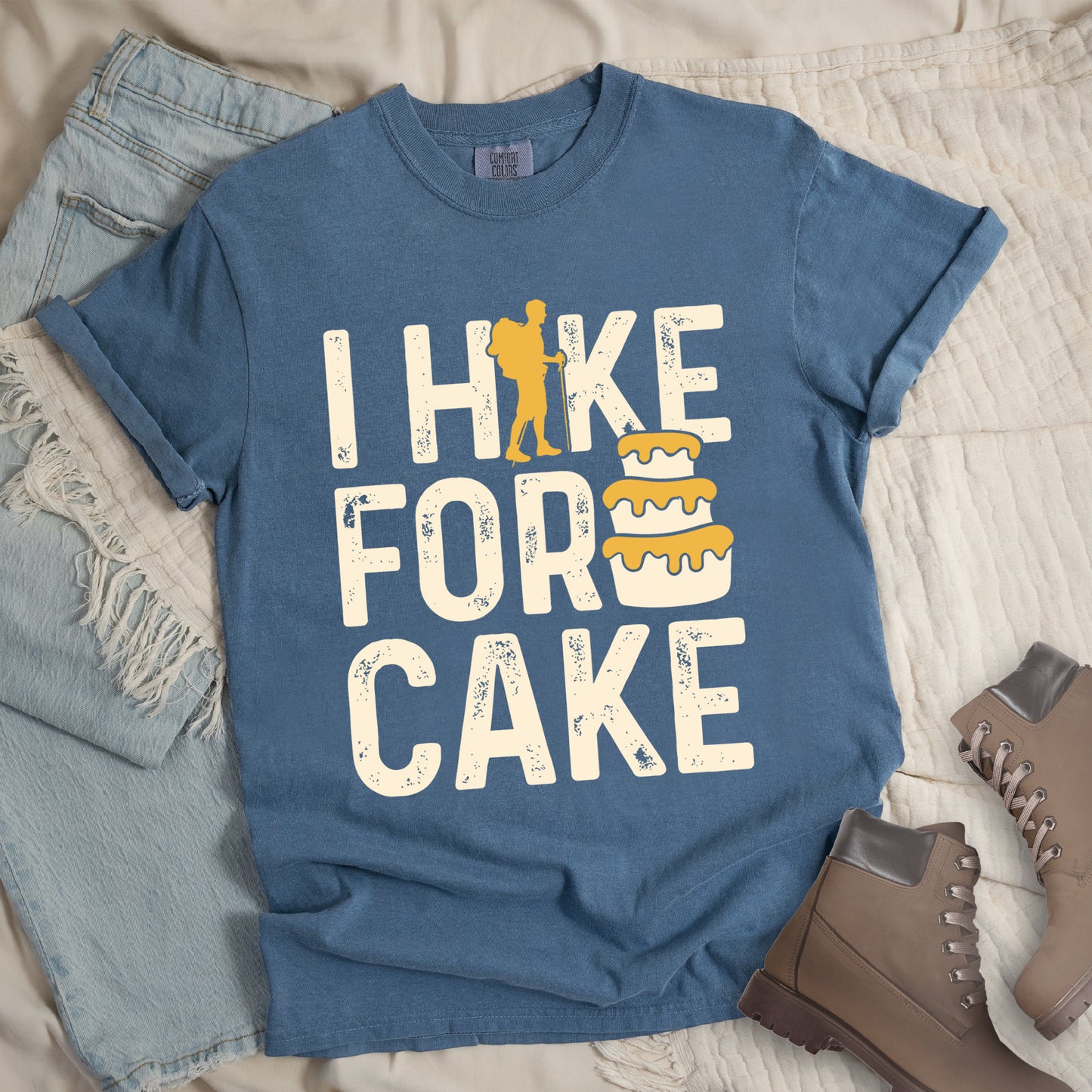 Blue Jean (Denim Blue) t-shirt with distressed cream-colored text reading "I HIKE FOR CAKE". A yellow hiker silhouette is cleverly incorporated into the letter "K," while a three-tiered cake with dripping yellow icing appears between "FOR" and "CAKE." The shirt features a classic crew neck and casual rolled sleeves, styled alongside light wash denim and brown hiking boots.
