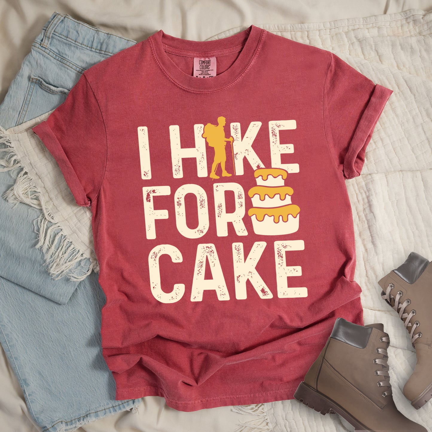 Crimson (Deep Pink) t-shirt with distressed cream-colored text reading "I HIKE FOR CAKE". A yellow hiker silhouette is cleverly incorporated into the letter "K," while a three-tiered cake with dripping yellow icing appears between "FOR" and "CAKE." The shirt features a classic crew neck and casual rolled sleeves, styled alongside light wash denim and brown hiking boots.
