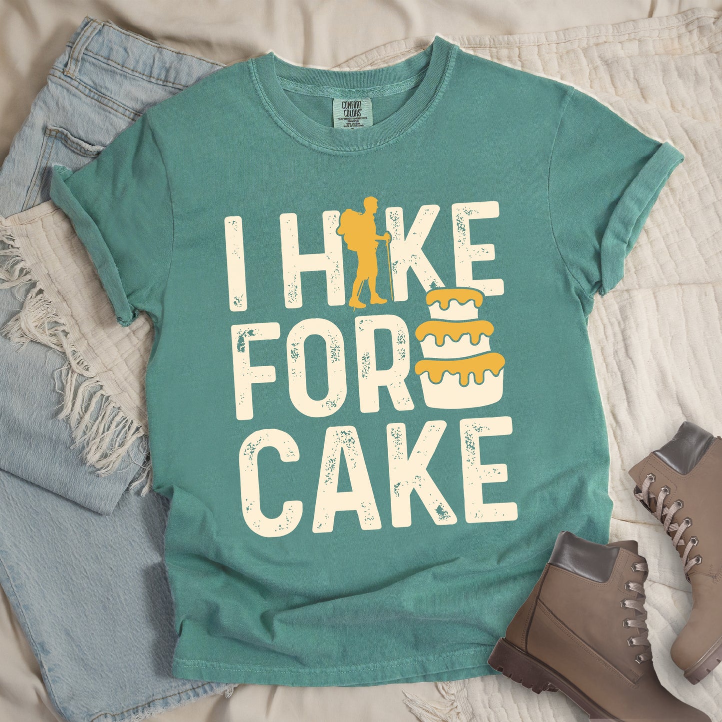 Light Green t-shirt with distressed cream-colored text reading "I HIKE FOR CAKE". A yellow hiker silhouette is cleverly incorporated into the letter "K," while a three-tiered cake with dripping yellow icing appears between "FOR" and "CAKE." The shirt features a classic crew neck and casual rolled sleeves, styled alongside light wash denim and brown hiking boots.