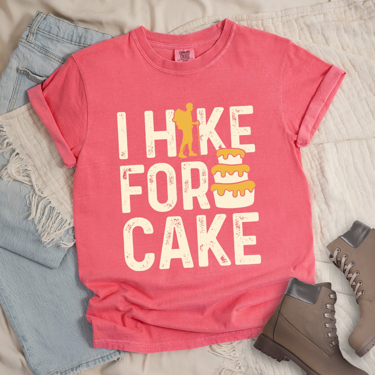 Watermelon (Light Coral) t-shirt displays "I HIKE FOR CAKE" in distressed off-white lettering. A yellow hiker silhouette is cleverly incorporated into the letter "K," while a three-tiered cake with dripping yellow icing appears between "FOR" and "CAKE." The shirt features a classic crew neck and casual rolled sleeves, styled alongside light wash denim and brown hiking boots.