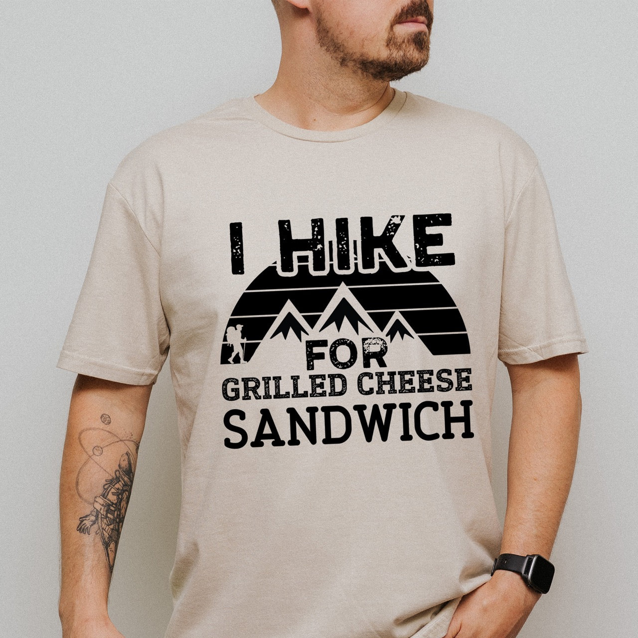 Man wearing sand (beige) I Hike for Grilled cheese Sandwich statement tee with bold distressed typography, geometric mountain design, and hiker silhouette for grilled cheese-loving outdoor enthusiasts, hikers and foodies