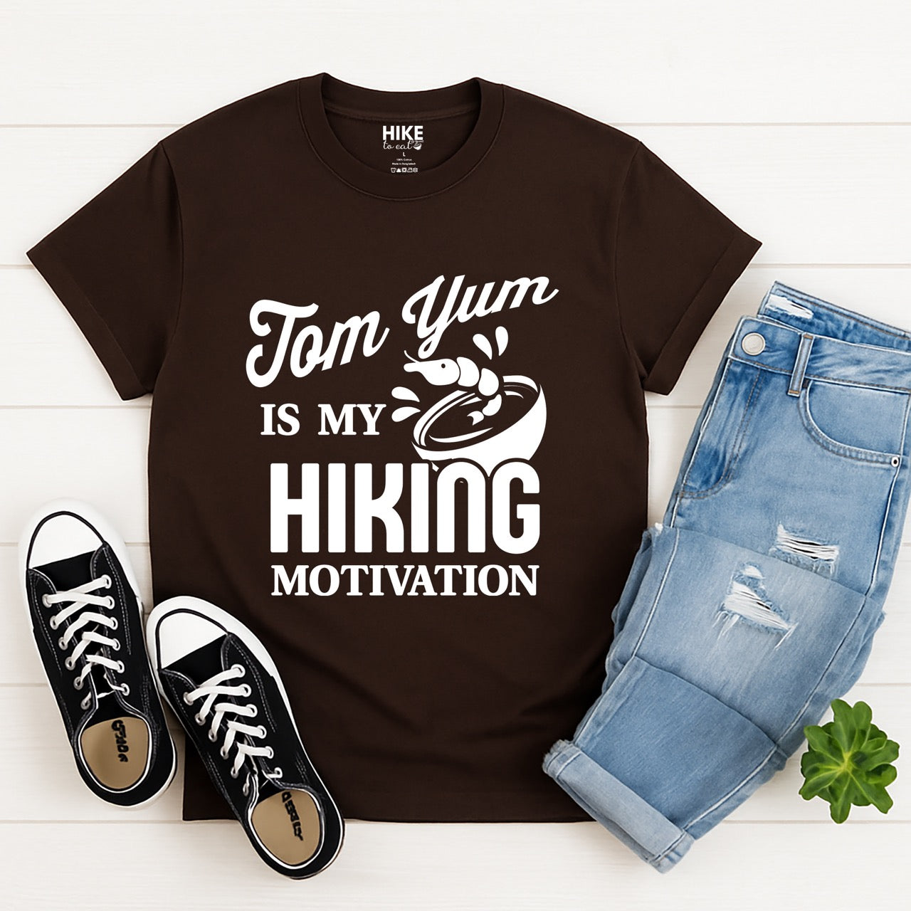 Chocolate brown Tom Yum is my Hiking Motivation statement tee with flowing script and bold typography, shrimp and soup bowl illustration for Thai food-loving outdoor enthusiasts, hikers and foodies laid flat with pants and shoes
