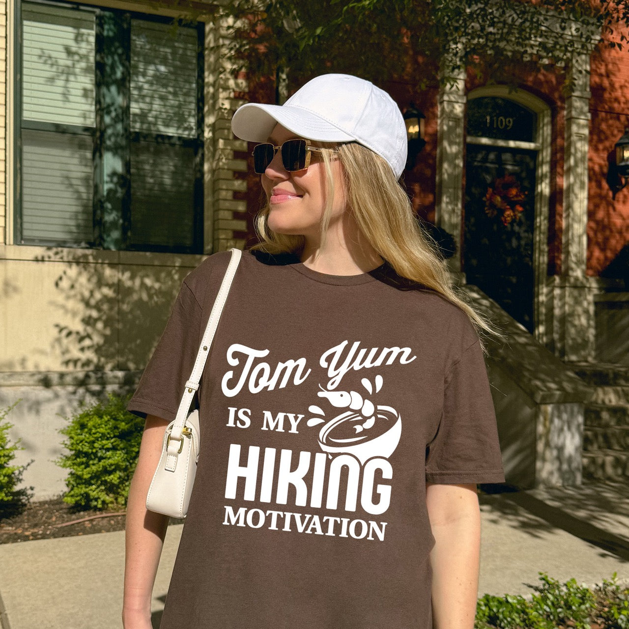 Woman wearing Chocolate brown Tom Yum is my Hiking Motivation statement tee with flowing script and bold typography, shrimp and soup bowl illustration for Thai food-loving outdoor enthusiasts, hikers and foodies