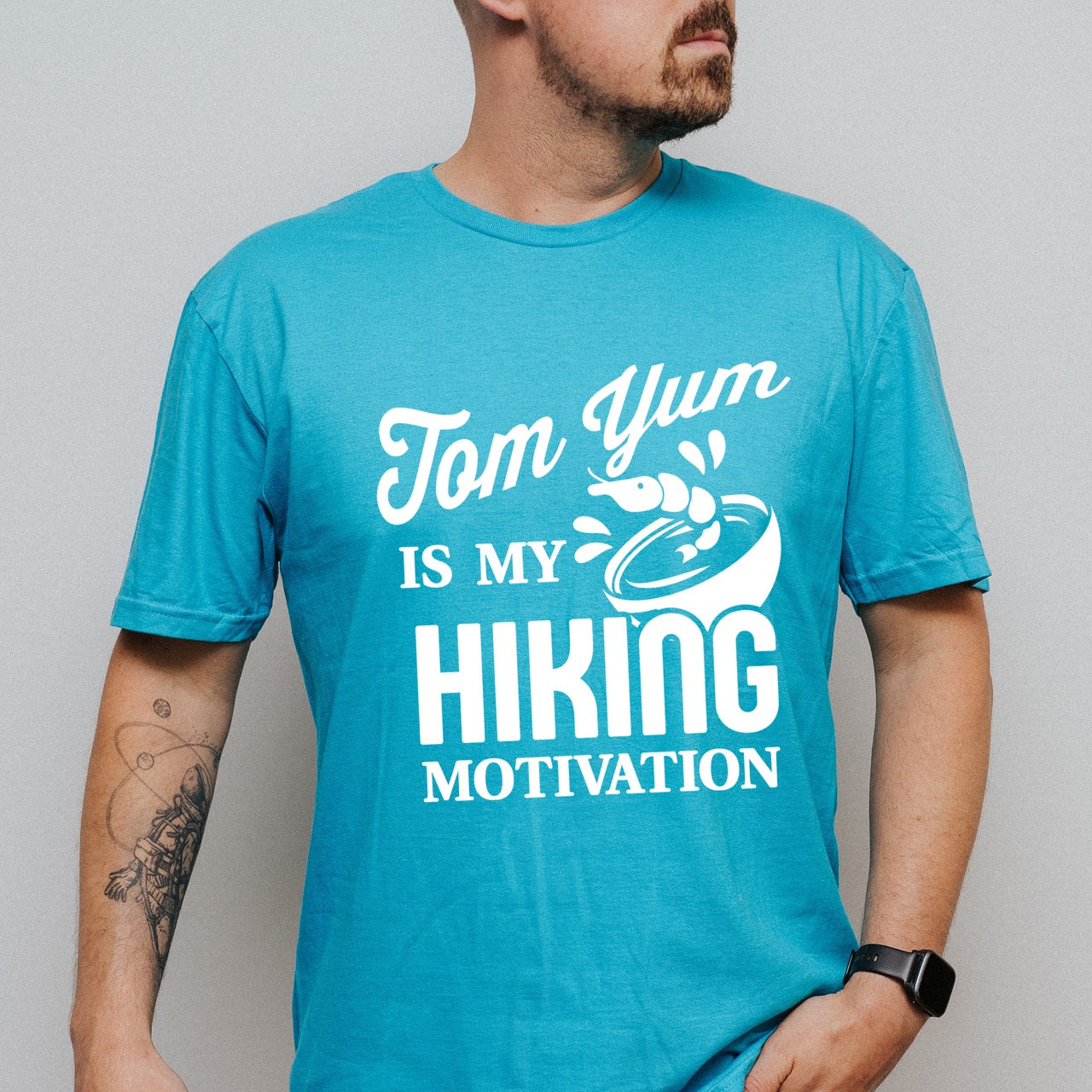 Man wearing Tropical blue Tom Yum is my Hiking Motivation statement tee with flowing script and bold typography, shrimp and soup bowl illustration for Thai food-loving outdoor enthusiasts, hikers and foodies