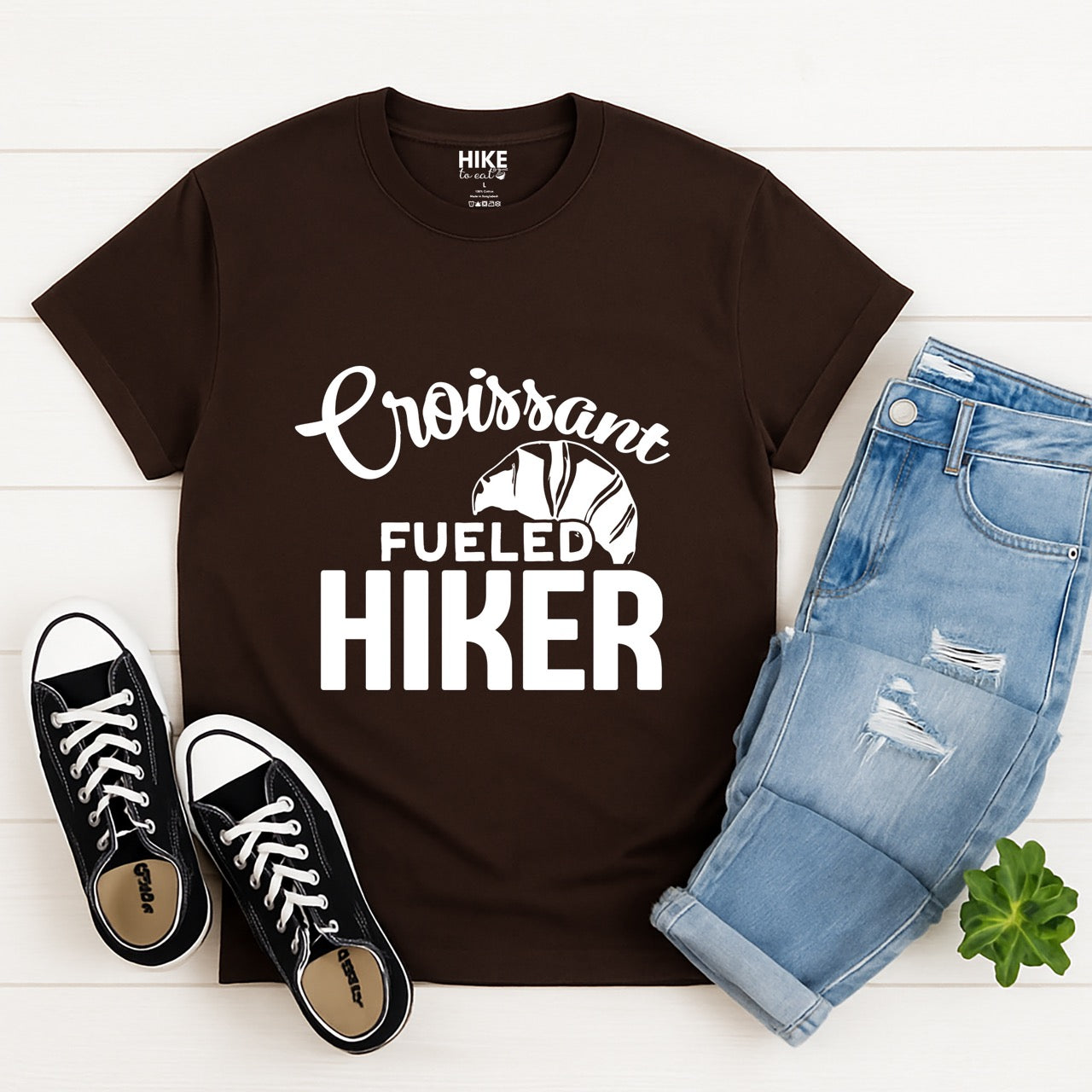 Chocolate brown Croissant Fueled Hiker statement tee with flowing script typography, bold lettering, and croissant illustration for French pastry-loving outdoor enthusiasts, hikers and foodies with pants and shoes