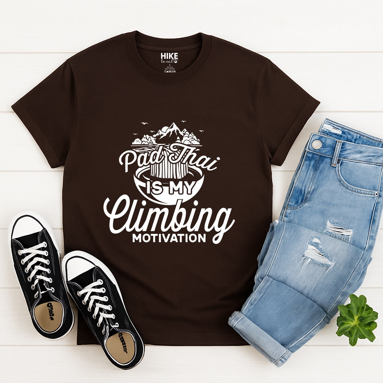 Dark chocolate Pad Thai is my Climbing motivation statement tee with flowing script typography, mountain landscape, noodle bowl illustration, and flying birds for Thai food-loving rock climber, hikers and foodies laid flat with pants and shoes