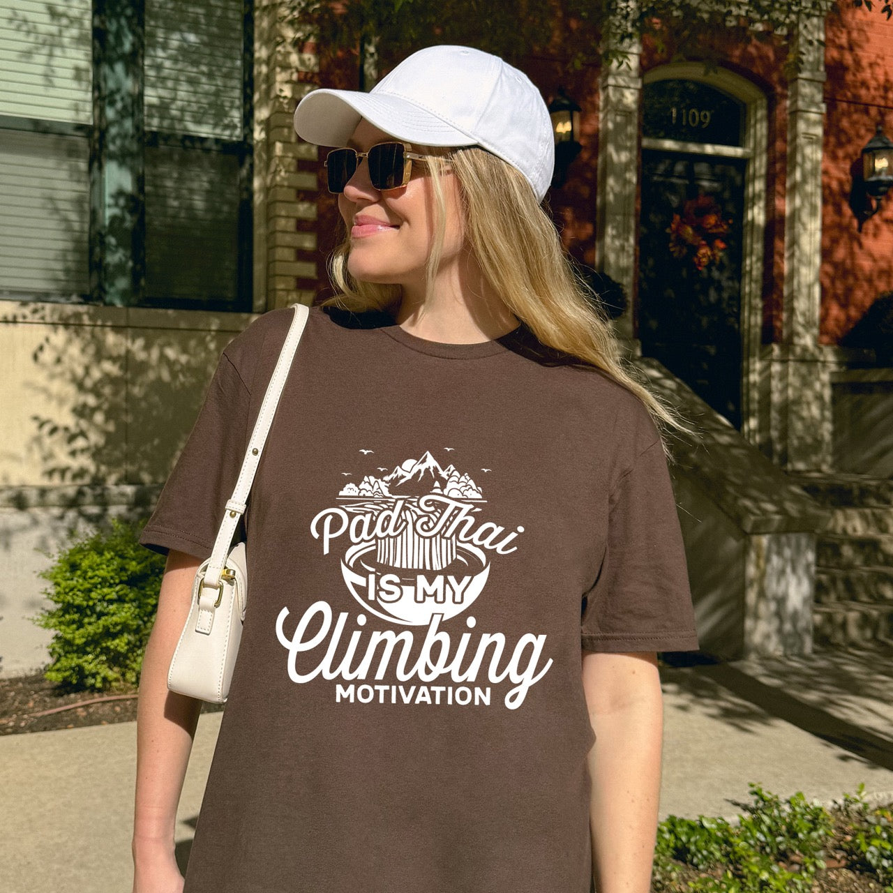 Woman wearing Dark chocolate Pad Thai is my Climbing motivation statement tee with flowing script typography, mountain landscape, noodle bowl illustration, and flying birds for Thai food-loving rock climber, hikers and foodies