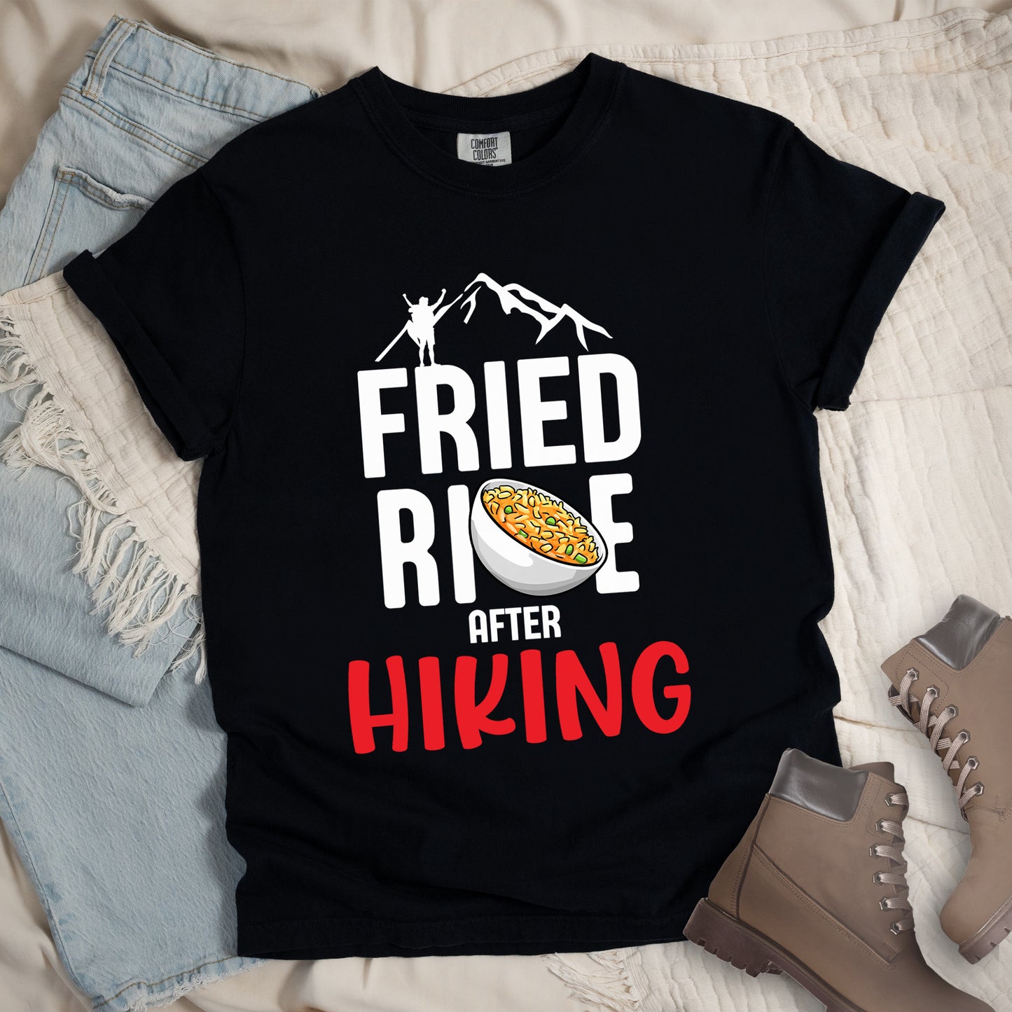 Black t-shirt with text "FRIED RICE AFTER HIKING". The top features white mountain silhouettes with a small hiker figure. "FRIED RICE" appears in large white letters with an illustration of a bowl of fried rice replacing the letter "C" in "RICE", tilted slightly to the right. The word "AFTER" is in small white text, and "HIKING" appears at the bottom in bold red letters.