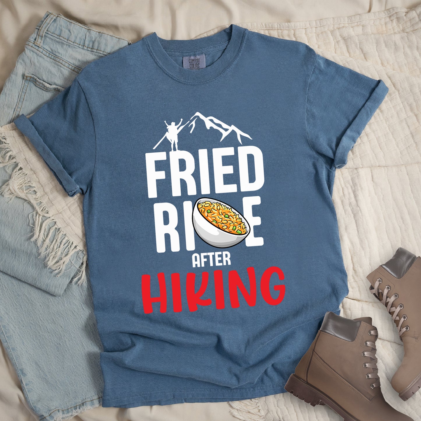 Blue Jean (Denim Blue) t-shirt with text "FRIED RICE AFTER HIKING". The top features white mountain silhouettes with a small hiker figure. "FRIED RICE" appears in large white letters with an illustration of a bowl of fried rice replacing the letter "C" in "RICE", tilted slightly to the right. The word "AFTER" is in small white text, and "HIKING" appears at the bottom in bold red letters.