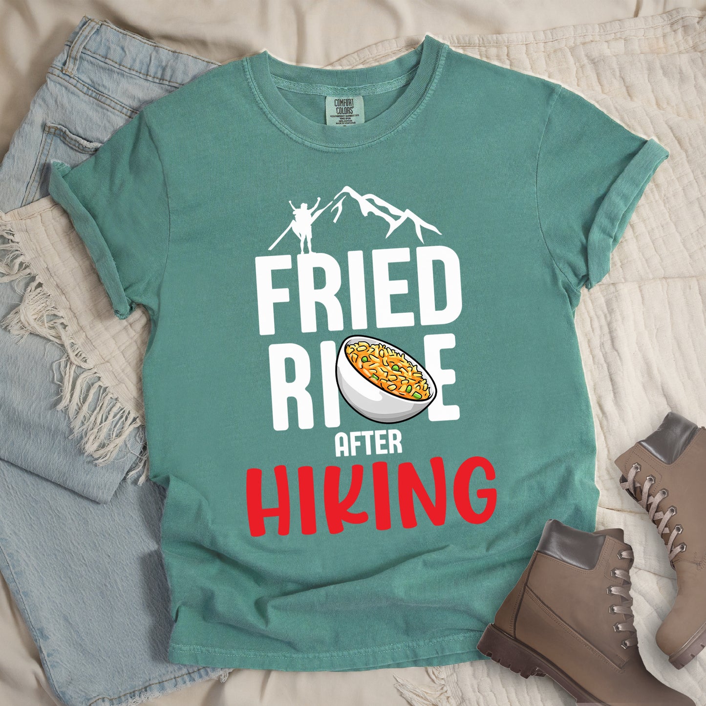 Light Green t-shirt with text "FRIED RICE AFTER HIKING". The top features white mountain silhouettes with a small hiker figure. "FRIED RICE" appears in large white letters with an illustration of a bowl of fried rice replacing the letter "C" in "RICE", tilted slightly to the right. The word "AFTER" is in small white text, and "HIKING" appears at the bottom in bold red letters.
