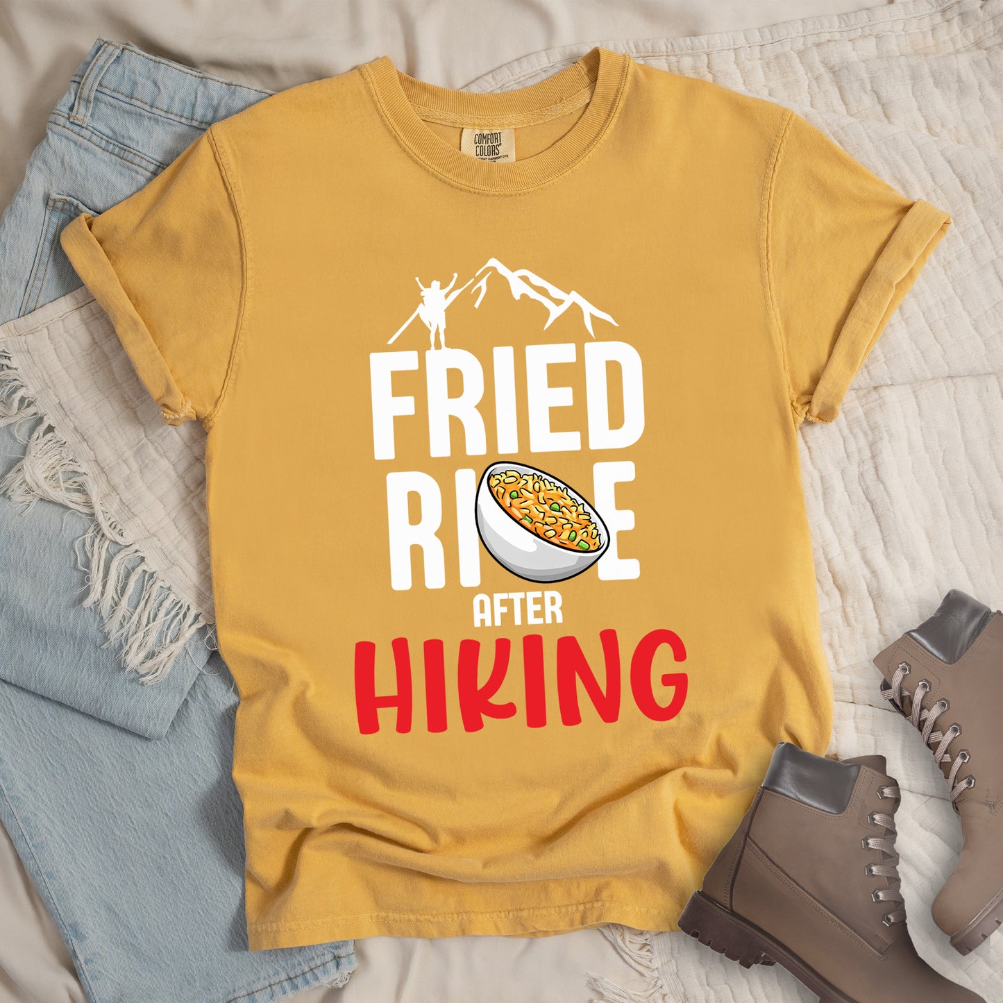 Mustard (Yellow) t-shirt with text "FRIED RICE AFTER HIKING". The top features white mountain silhouettes with a small hiker figure. "FRIED RICE" appears in large white letters with an illustration of a bowl of fried rice replacing the letter "C" in "RICE", tilted slightly to the right. The word "AFTER" is in small white text, and "HIKING" appears at the bottom in bold red letters.