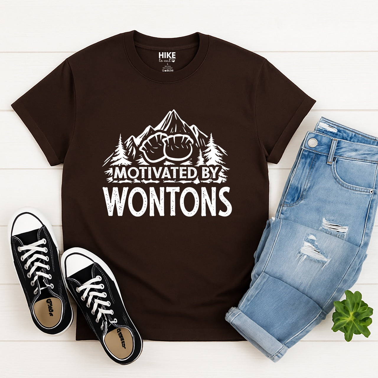 Dark chocolate Motivated by Wontons statement tee with bold distressed typography, mountain peaks, pine trees, and wonton illustration for Chinese food-loving outdoor enthusiasts, hikers and foodies laid flat with shoes and pants