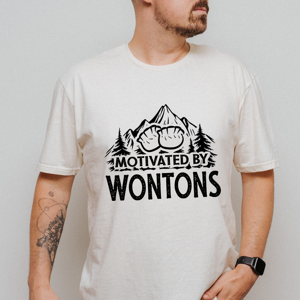 Man wearing Natural (beige) Motivated by Wontons statement tee with bold distressed typography, mountain peaks, pine trees, and wonton illustration for Chinese food-loving outdoor enthusiasts, hikers and foodies
