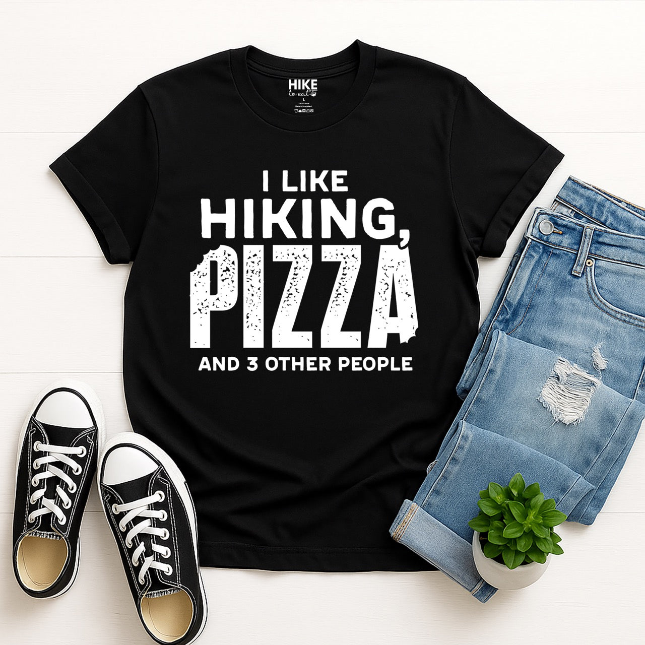 Black Motivated by Wontons statement tee with bold distressed typography, mountain peaks, pine trees, and wonton illustration for Chinese food-loving outdoor enthusiasts, hikers and foodies laid flat with pants and shoes
