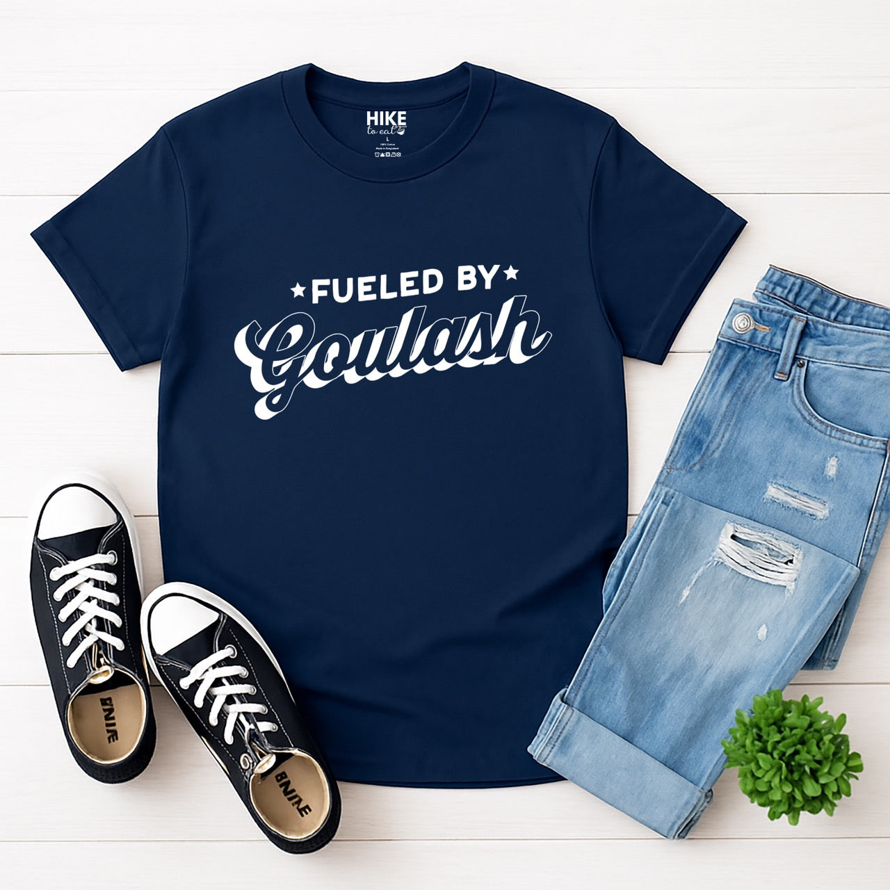 Navy Hungarian food lover Fueled by Goulash statement shirt with bold distressed lettering and flowing script design featuring decorative stars for outdoor adventurers who love paprika-spiced comfort stew for hikers, climbers and foodies laid flat with pants and shoes