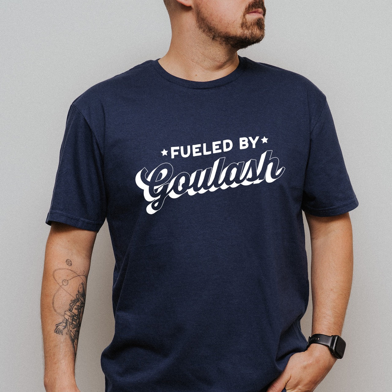 Man wearing Navy Hungarian food lover Fueled by Goulash statement shirt with bold distressed lettering and flowing script design featuring decorative stars for outdoor adventurers who love paprika-spiced comfort stew for hikers, climbers and foodies