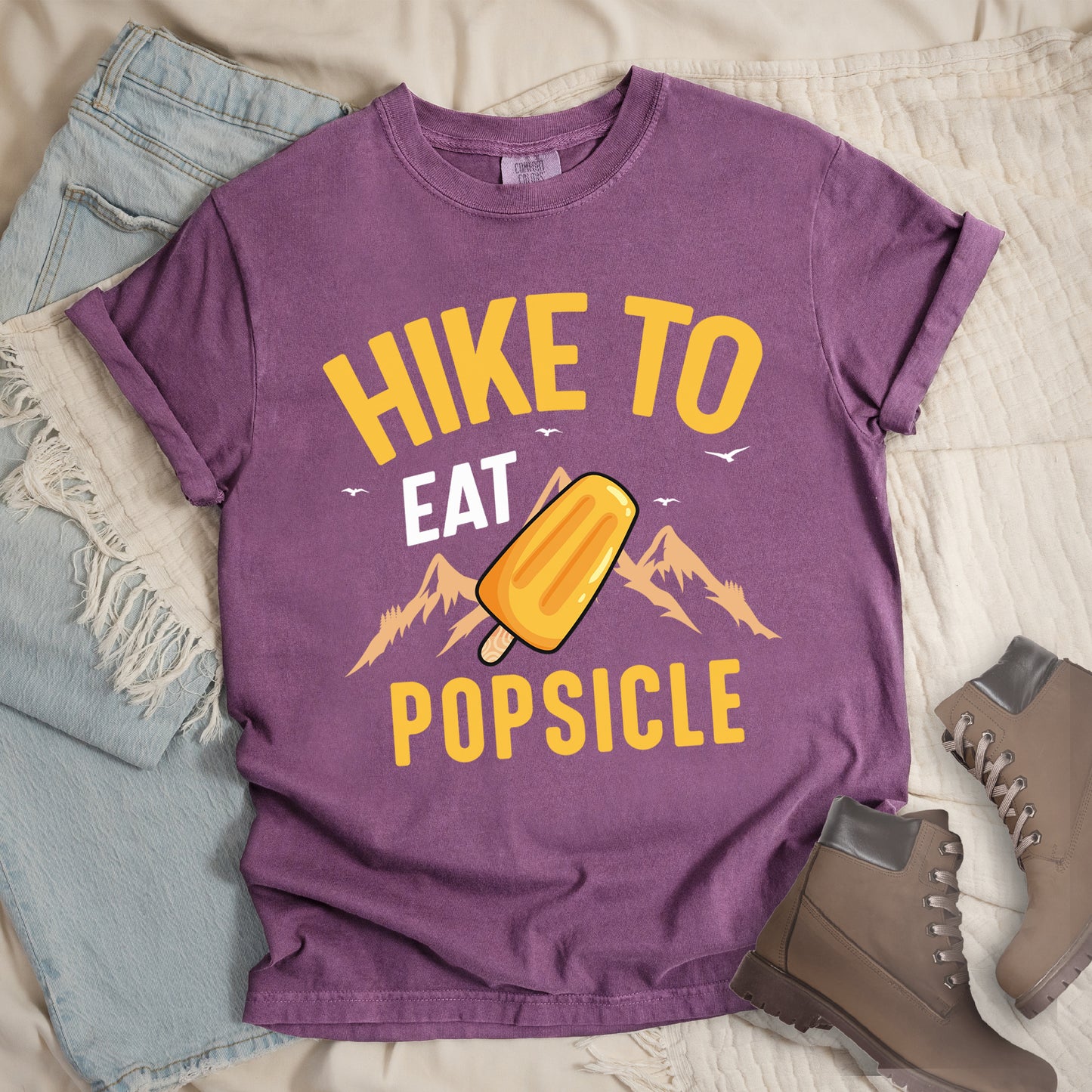 Berry (Dark Violet) t-shirt with text "HIKE TO EAT POPSICLE". "HIKE TO" appears in yellow at the top, "EAT" in white in the middle, and "POPSICLE" in yellow at the bottom. Between "EAT" and "POPSICLE" is an illustration of an orange popsicle on a stick positioned in front of light orange mountain silhouettes. Small white bird silhouettes appear near the top of the design. Features a classic crew neck and casual rolled sleeves, styled alongside light wash denim and brown hiking boots.