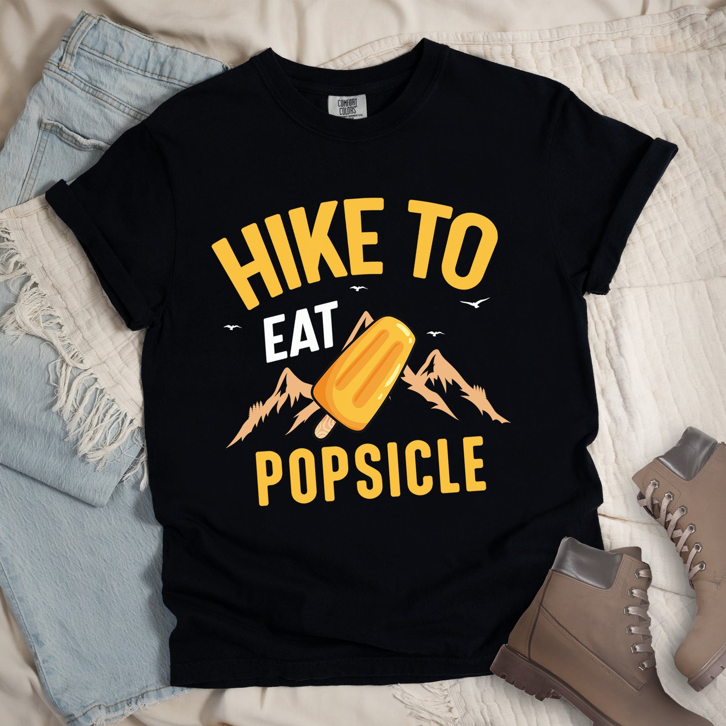 Black t-shirt with text "HIKE TO EAT POPSICLE". "HIKE TO" appears in yellow at the top, "EAT" in white in the middle, and "POPSICLE" in yellow at the bottom. Between "EAT" and "POPSICLE" is an illustration of an orange popsicle on a stick positioned in front of light orange mountain silhouettes. Small white bird silhouettes appear near the top of the design. Features a classic crew neck and casual rolled sleeves, styled alongside light wash denim and brown hiking boots.