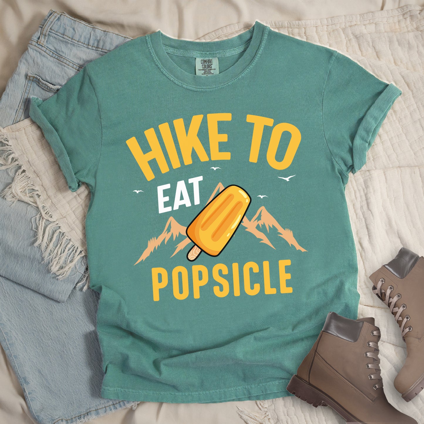 Light Green t-shirt with text "HIKE TO EAT POPSICLE". "HIKE TO" appears in yellow at the top, "EAT" in white in the middle, and "POPSICLE" in yellow at the bottom. Between "EAT" and "POPSICLE" is an illustration of an orange popsicle on a stick positioned in front of light orange mountain silhouettes. Small white bird silhouettes appear near the top of the design. Features a classic crew neck and casual rolled sleeves, styled alongside light wash denim and brown hiking boots.