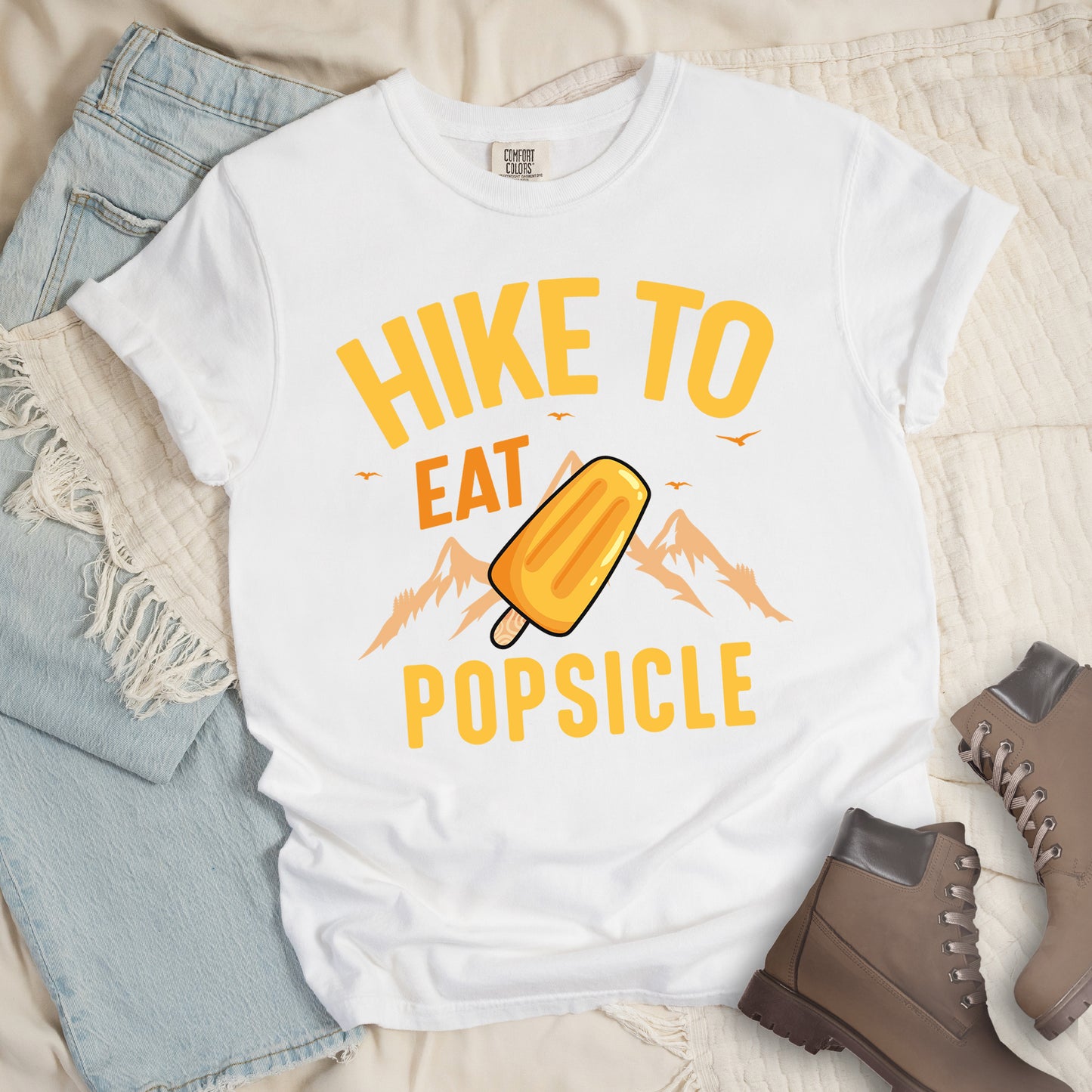 White t-shirt with text "HIKE TO EAT POPSICLE". "HIKE TO" appears in yellow at the top, "EAT" in orange in the middle, and "POPSICLE" in yellow at the bottom. Between "EAT" and "POPSICLE" is an illustration of an orange popsicle on a stick positioned in front of light orange mountain silhouettes. Small orange bird silhouettes appear near the top of the design. Features a classic crew neck and casual rolled sleeves, styled alongside light wash denim and brown hiking boots.