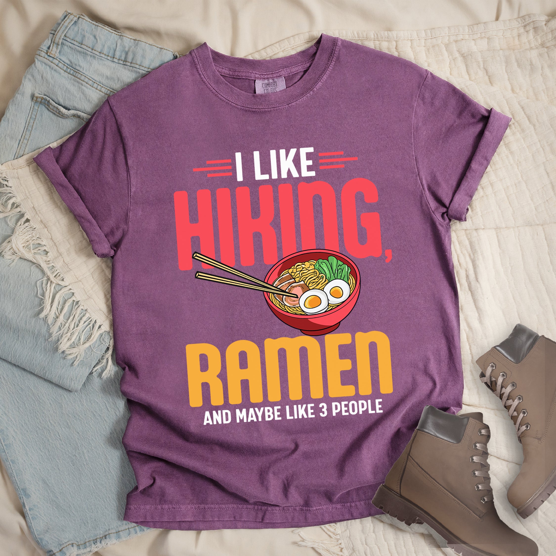 Berry (Dark Violet) t-shirt with text "I LIKE HIKING, RAMEN AND MAYBE LIKE 3 PEOPLE". "I LIKE" appears in white at the top with red decorative lines, "HIKING," in large red letters, "RAMEN" in yellow/orange, and "AND MAYBE LIKE 3 PEOPLE" in white at the bottom. Between "HIKING" and "RAMEN" is a colorful illustration of a red ramen bowl with noodles, egg, and vegetables, with chopsticks. The shirt features a classic crew neck and casual rolled sleeves, styled alongside light wash denim and brown hiking boots