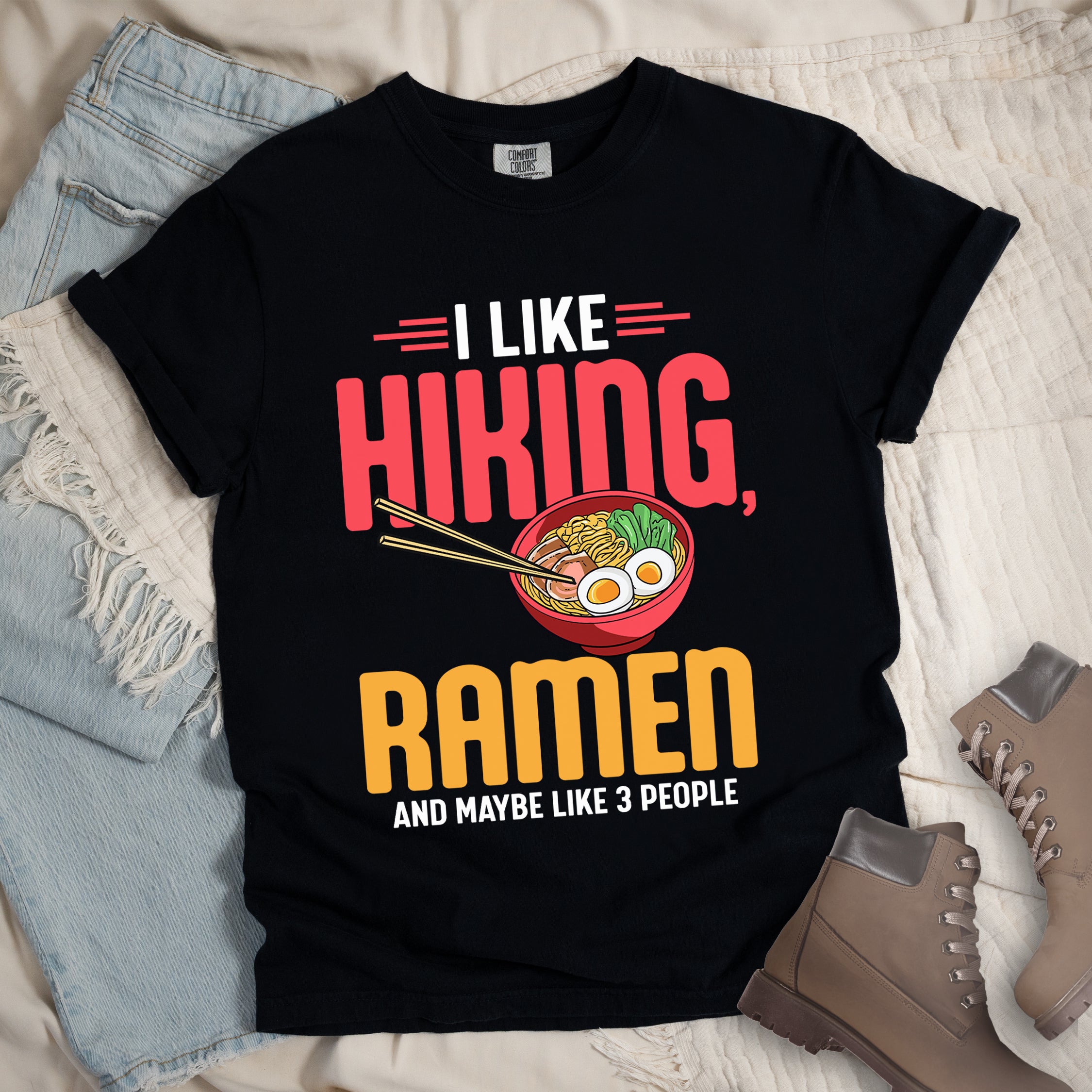 Black t-shirt with text "I LIKE HIKING, RAMEN AND MAYBE LIKE 3 PEOPLE". "I LIKE" appears in white at the top with red decorative lines, "HIKING," in large red letters, "RAMEN" in yellow/orange, and "AND MAYBE LIKE 3 PEOPLE" in white at the bottom. Between "HIKING" and "RAMEN" is a colorful illustration of a red ramen bowl with noodles, egg, and vegetables, with chopsticks. The shirt features a classic crew neck and casual rolled sleeves, styled alongside light wash denim and brown hiking boots.
