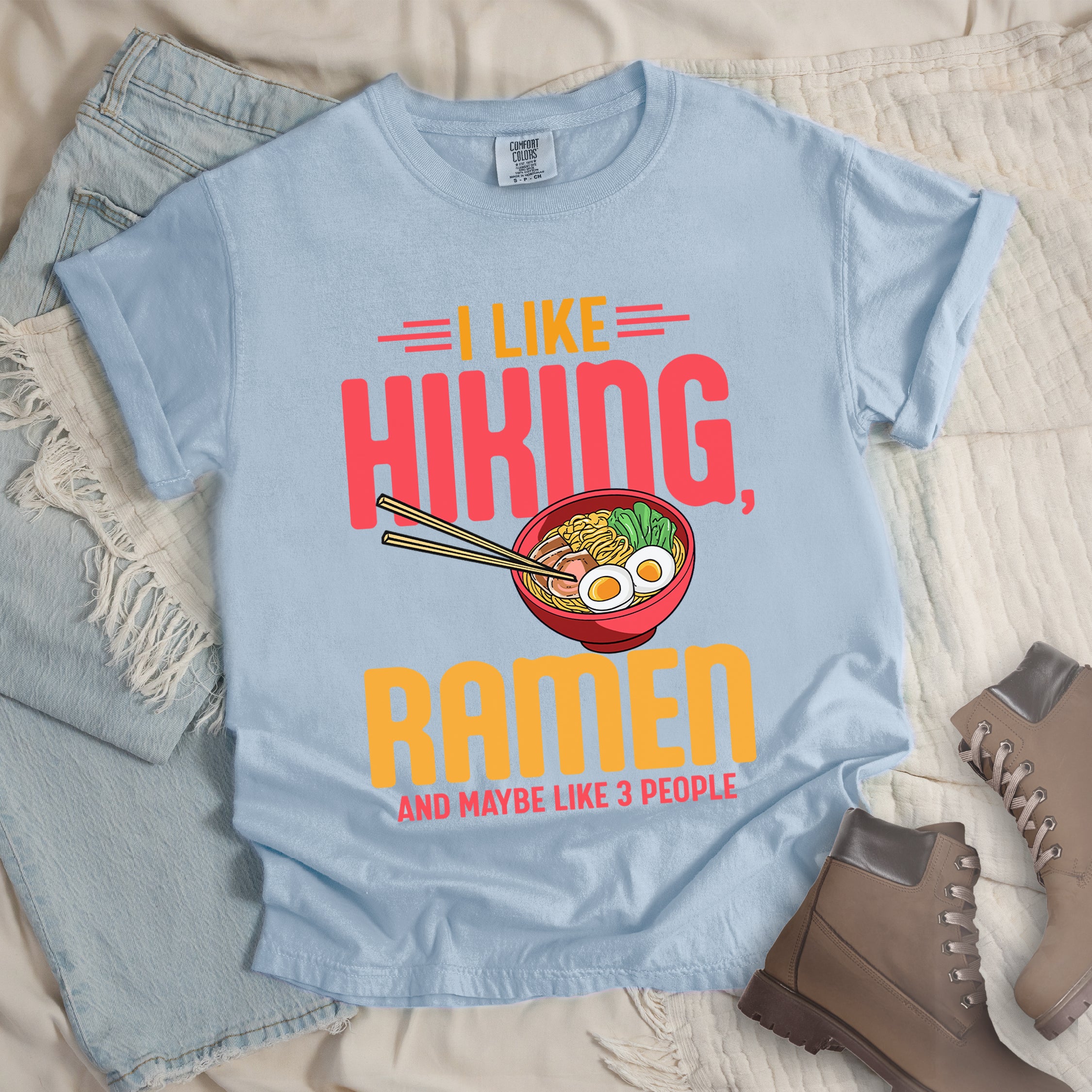 Chambray (Light Blue) t-shirt with text "I LIKE HIKING, RAMEN AND MAYBE LIKE 3 PEOPLE". "I LIKE" appears in orange at the top with red decorative lines, "HIKING," in large red letters, "RAMEN" in yellow/orange, and "AND MAYBE LIKE 3 PEOPLE" in red at the bottom. Between "HIKING" and "RAMEN" is a colorful illustration of a red ramen bowl with noodles, egg, and vegetables, with chopsticks. Features a classic crew neck and casual rolled sleeves, styled alongside light wash denim and brown hiking boots.