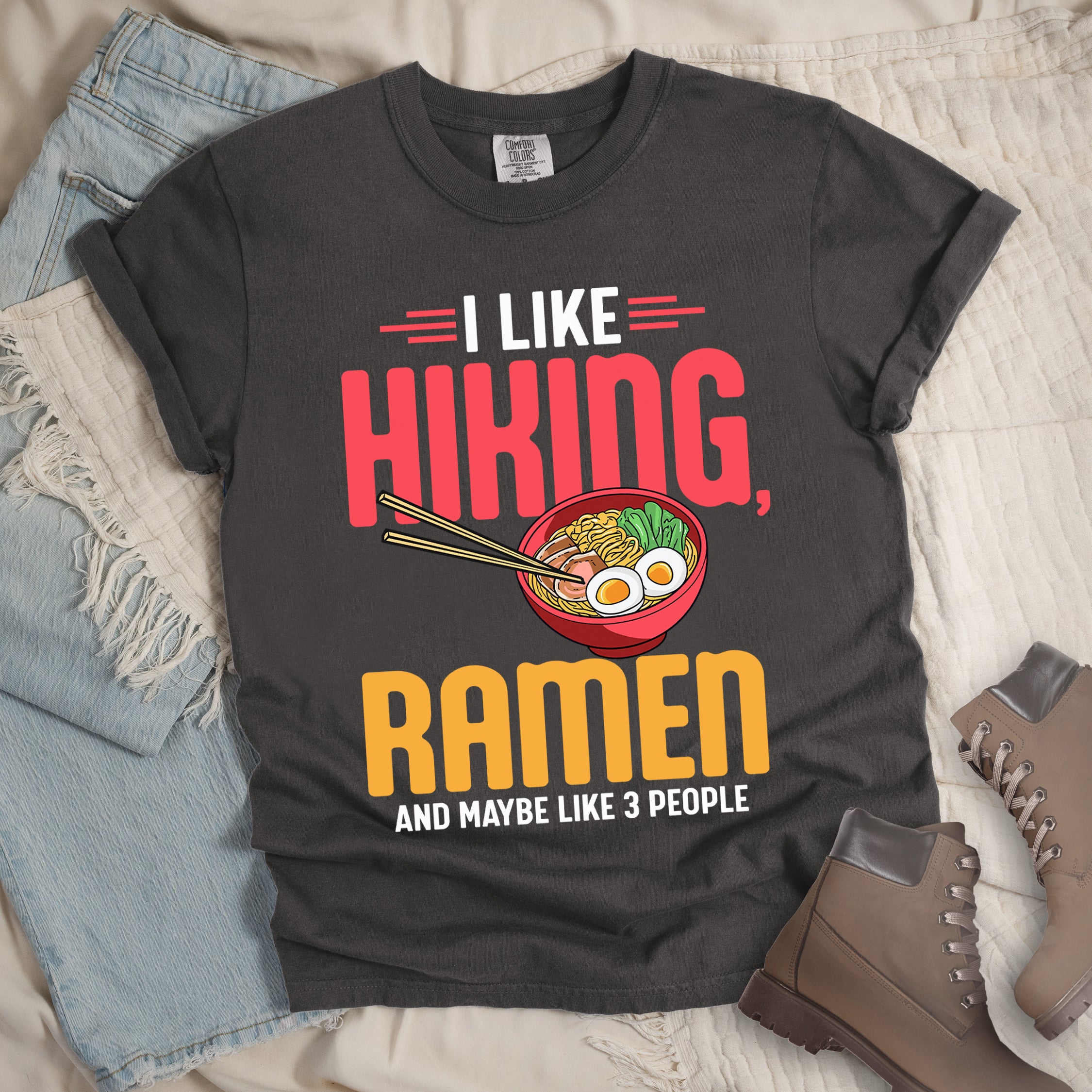Pepper (Dark Gray) t-shirt with text "I LIKE HIKING, RAMEN AND MAYBE LIKE 3 PEOPLE". "I LIKE" appears in white at the top with red decorative lines, "HIKING," in large red letters, "RAMEN" in yellow/orange, and "AND MAYBE LIKE 3 PEOPLE" in white at the bottom. Between "HIKING" and "RAMEN" is a colorful illustration of a red ramen bowl with noodles, egg, and vegetables, with chopsticks. Features a classic crew neck and casual rolled sleeves, styled alongside light wash denim and brown hiking boots.