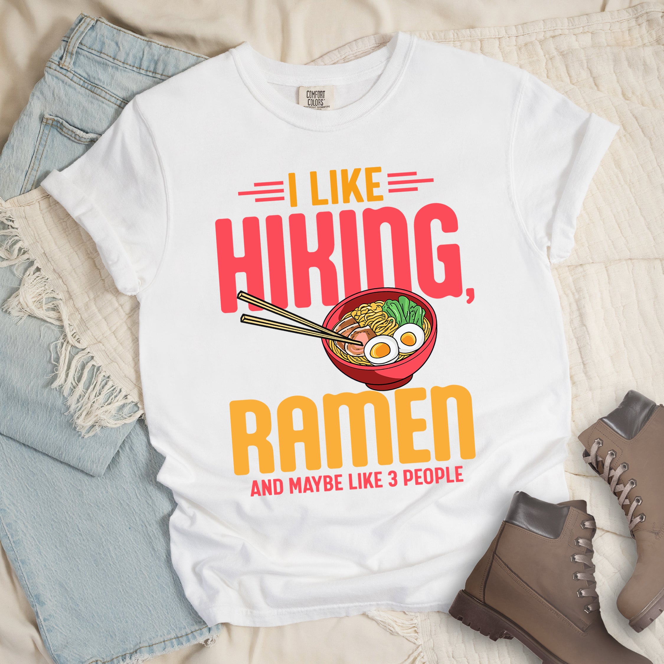 White t-shirt with text "I LIKE HIKING, RAMEN AND MAYBE LIKE 3 PEOPLE". "I LIKE" appears in orange at the top with red decorative lines, "HIKING," in large red letters, "RAMEN" in yellow/orange, and "AND MAYBE LIKE 3 PEOPLE" in red at the bottom. Between "HIKING" and "RAMEN" is a colorful illustration of a red ramen bowl with noodles, egg, and vegetables, with chopsticks. The shirt features a classic crew neck and casual rolled sleeves, styled alongside light wash denim and brown hiking boots.