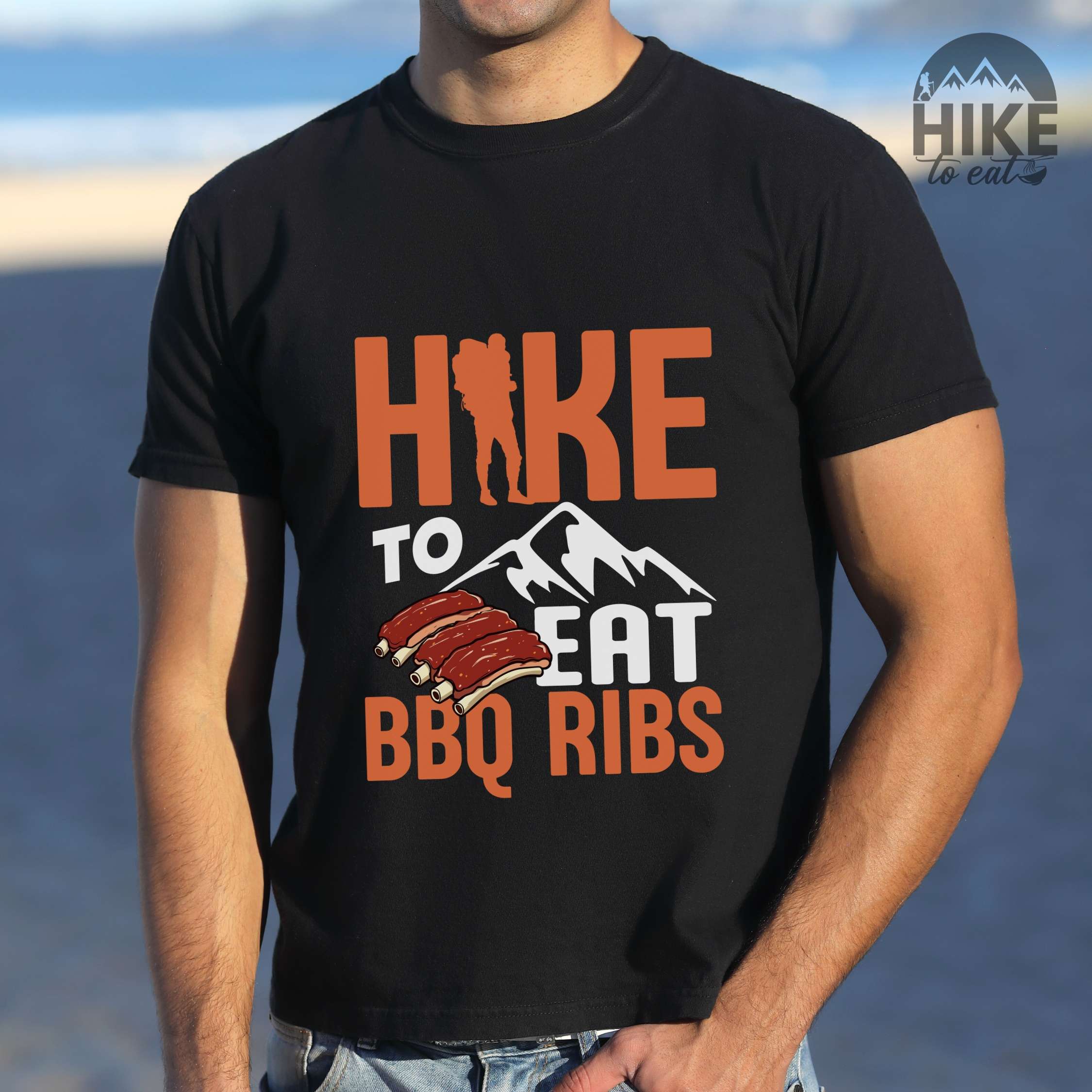 Man wearing black "Hike to eat BBQ ribs" colored vintage tee with bold typography, mountains, juicy grilled bbq ribs illustration, and backpacker for bbq enthusiasts