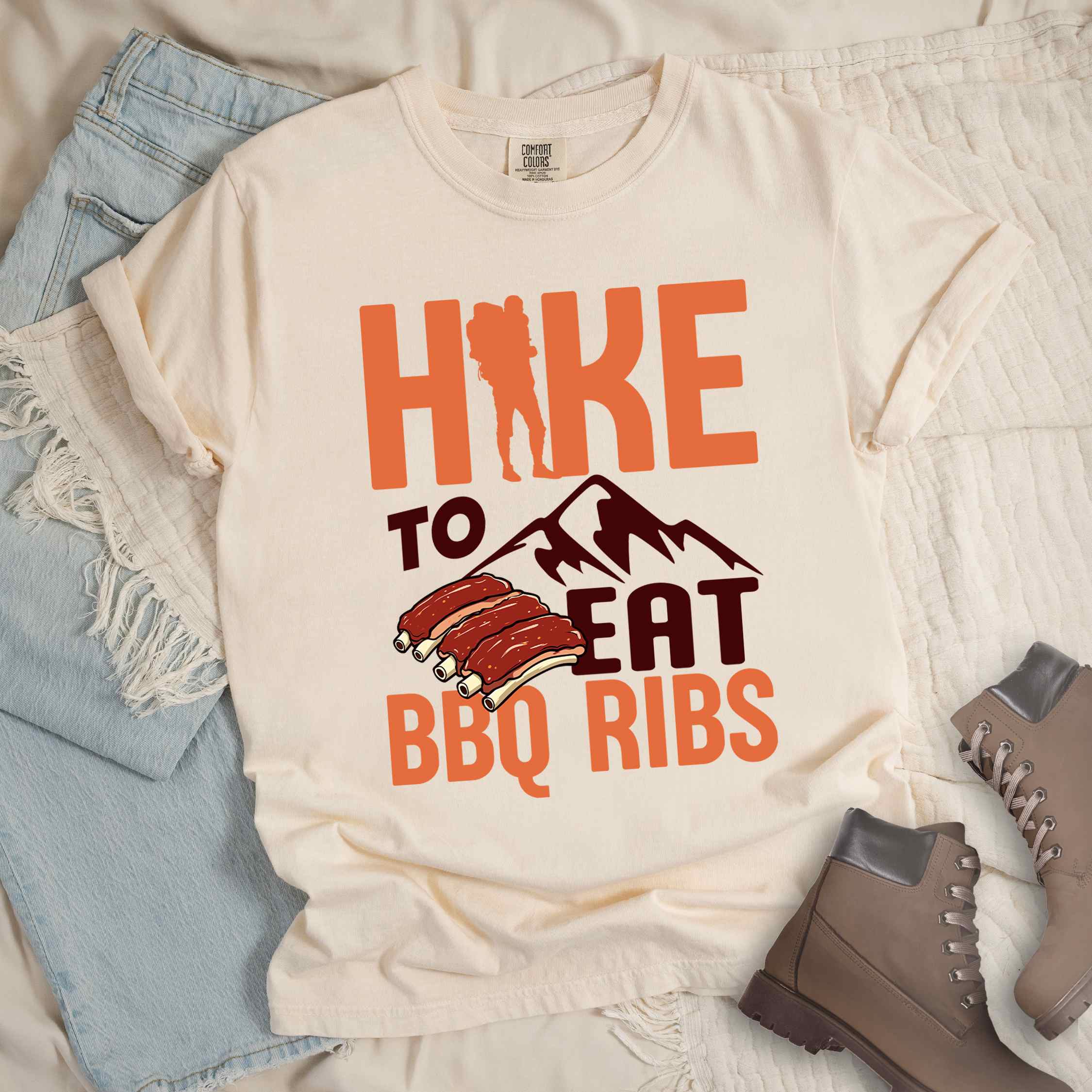 Ivory "Hike to eat BBQ ribs" colored vintage tee with bold typography, mountains, juicy grilled bbq ribs illustration, and backpacker for bbq enthusiasts