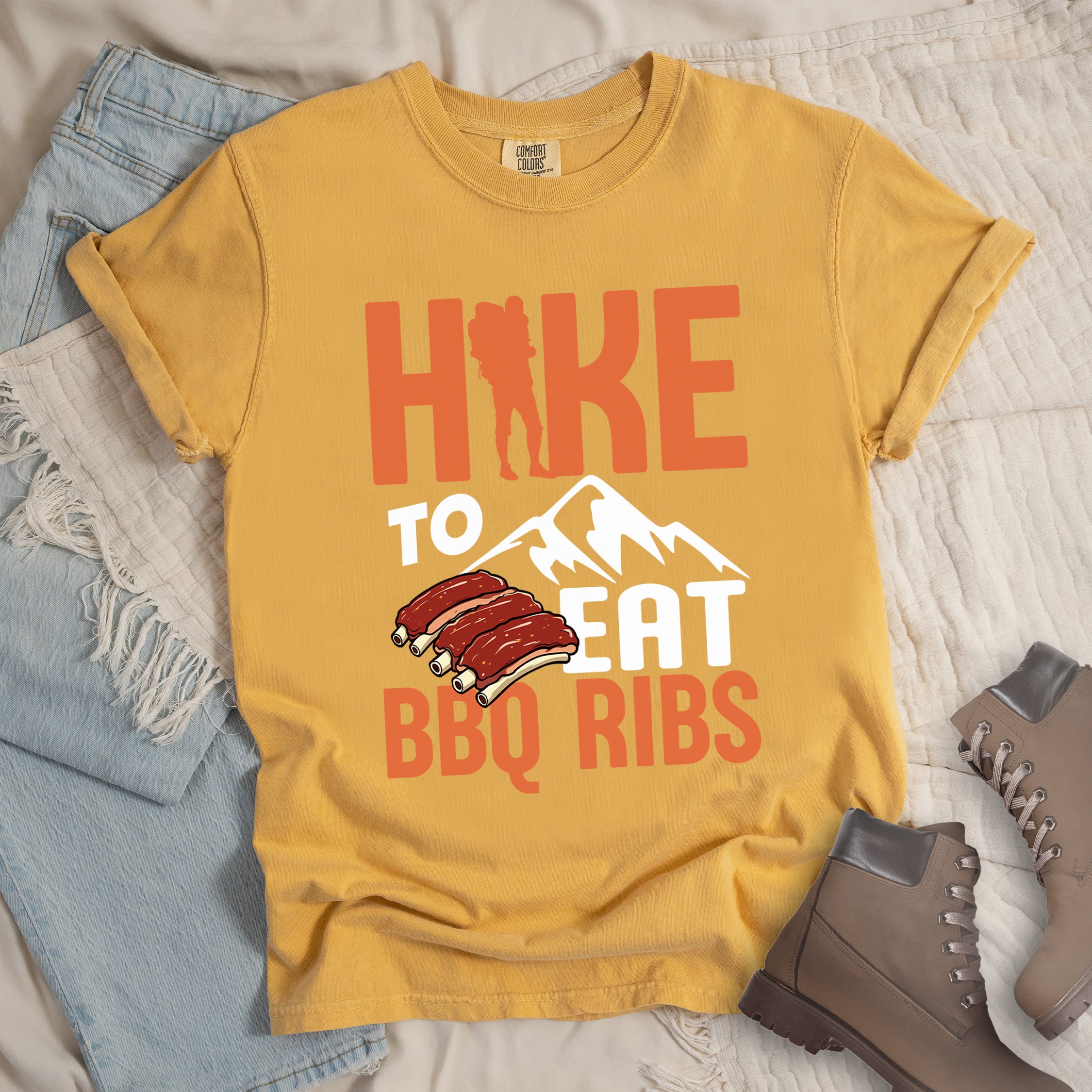 Mustard (Yellow) t-shirt with text "HIKE TO EAT BBQ RIBS". The word "HIKE" appears in orange with a hiker silhouette replacing the "I". Below that is "TO" in white text above a white mountain graphic. "EAT" appears in white, and "BBQ RIBS" in orange at the bottom. Between "EAT" and "BBQ RIBS" is an illustration of a rack of barbecue ribs with exposed bones.