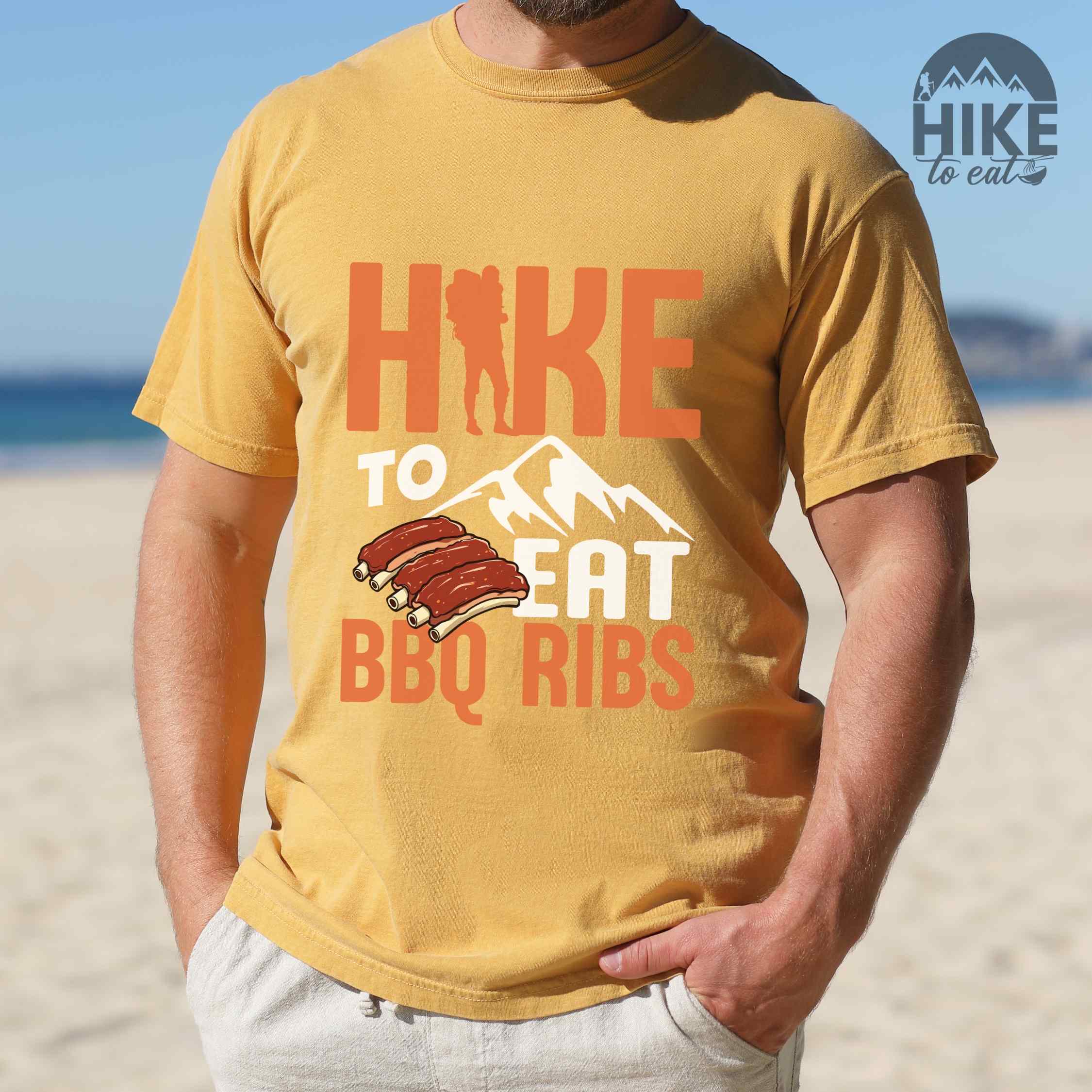 Man wearing mustard "Hike to eat BBQ ribs" colored vintage tee with bold typography, mountains, juicy grilled bbq ribs illustration, and backpacker for bbq enthusiasts