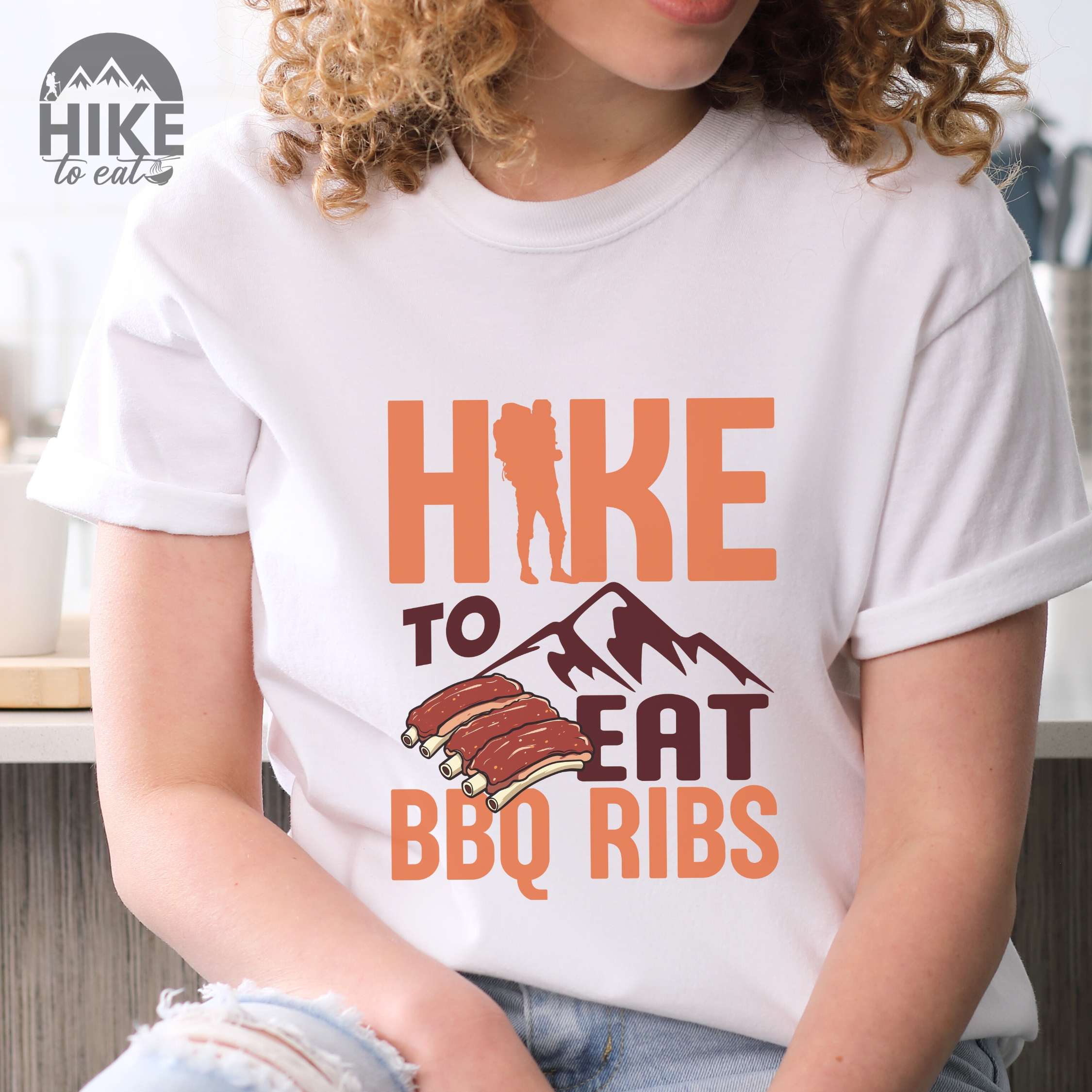 Woman wearing white "Hike to eat BBQ ribs" colored vintage tee with bold typography, mountains, juicy grilled bbq ribs illustration, and backpacker for bbq enthusiasts
