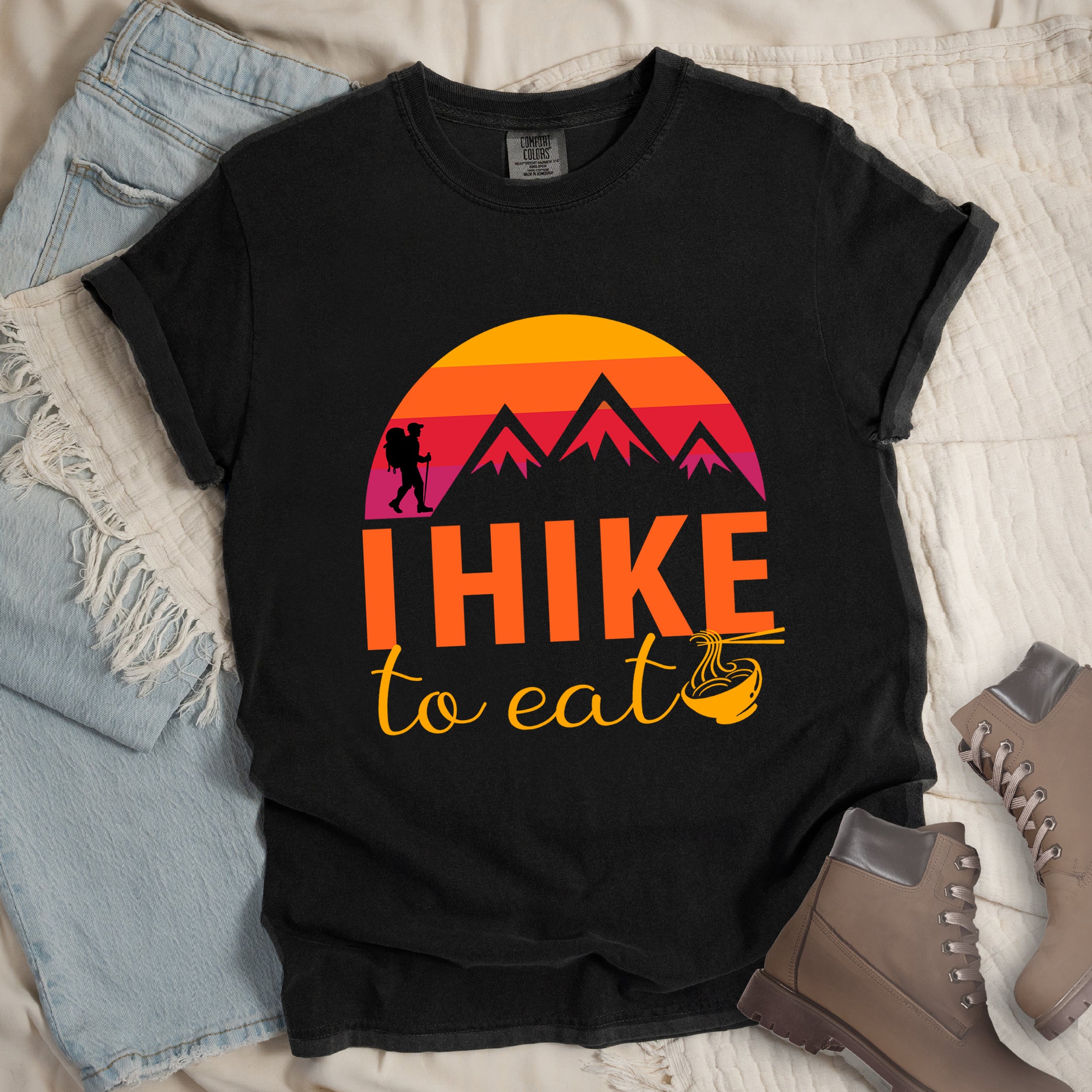 Black colored t-shirt featuring a retro sunset design with gradient colors from yellow to orange to red to purple. Mountain silhouettes and a small hiker figure appear against the sunset. Below this is "I HIKE" in large orange letters and "to eat" in yellow script, followed by a small illustration of a noodle bowl with chopsticks. The design has a vintage outdoor aesthetic. Features a classic crew neck and casual rolled sleeves, styled alongside light wash denim and brown hiking boots.