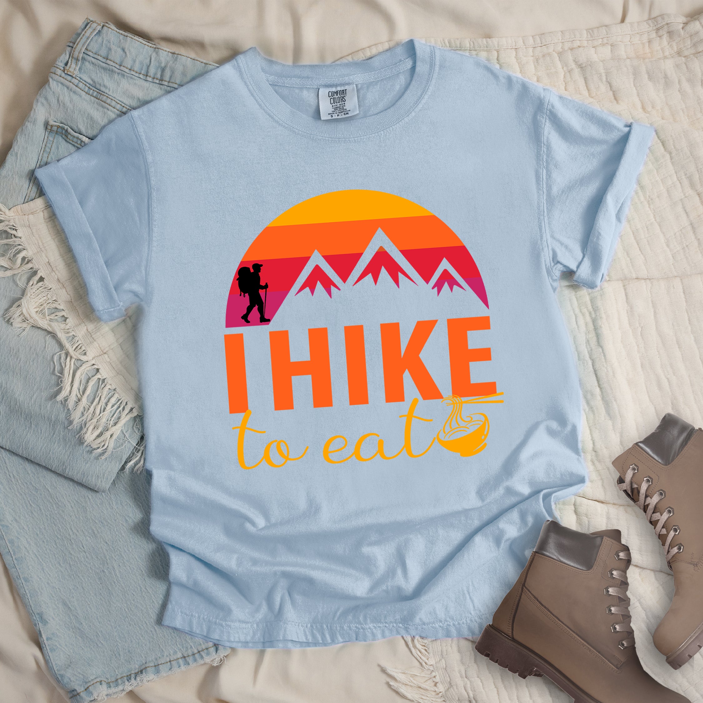 Chambray colored t-shirt featuring a retro sunset design with gradient colors from yellow to orange to red to purple. Mountain silhouettes and a small hiker figure appear against the sunset. Below this is "I HIKE" in large orange letters and "to eat" in yellow script, followed by a small illustration of a noodle bowl with chopsticks. The design has a vintage outdoor aesthetic. Features a classic crew neck and casual rolled sleeves, styled alongside light wash denim and brown hiking boots.