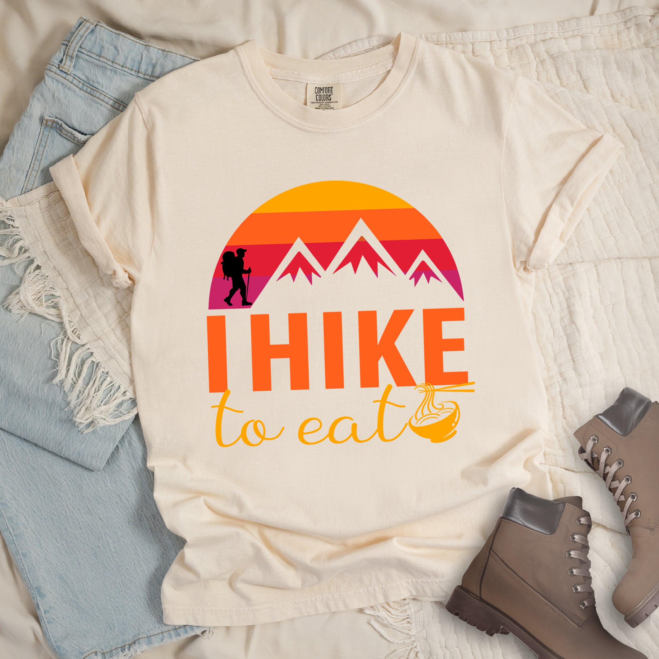 Ivory colored t-shirt featuring a retro sunset design with gradient colors from yellow to orange to red to purple. Mountain silhouettes and a small hiker figure appear against the sunset. Below this is "I HIKE" in large orange letters and "to eat" in yellow script, followed by a small illustration of a noodle bowl with chopsticks. The design has a vintage outdoor aesthetic. Features a classic crew neck and casual rolled sleeves, styled alongside light wash denim and brown hiking boots.