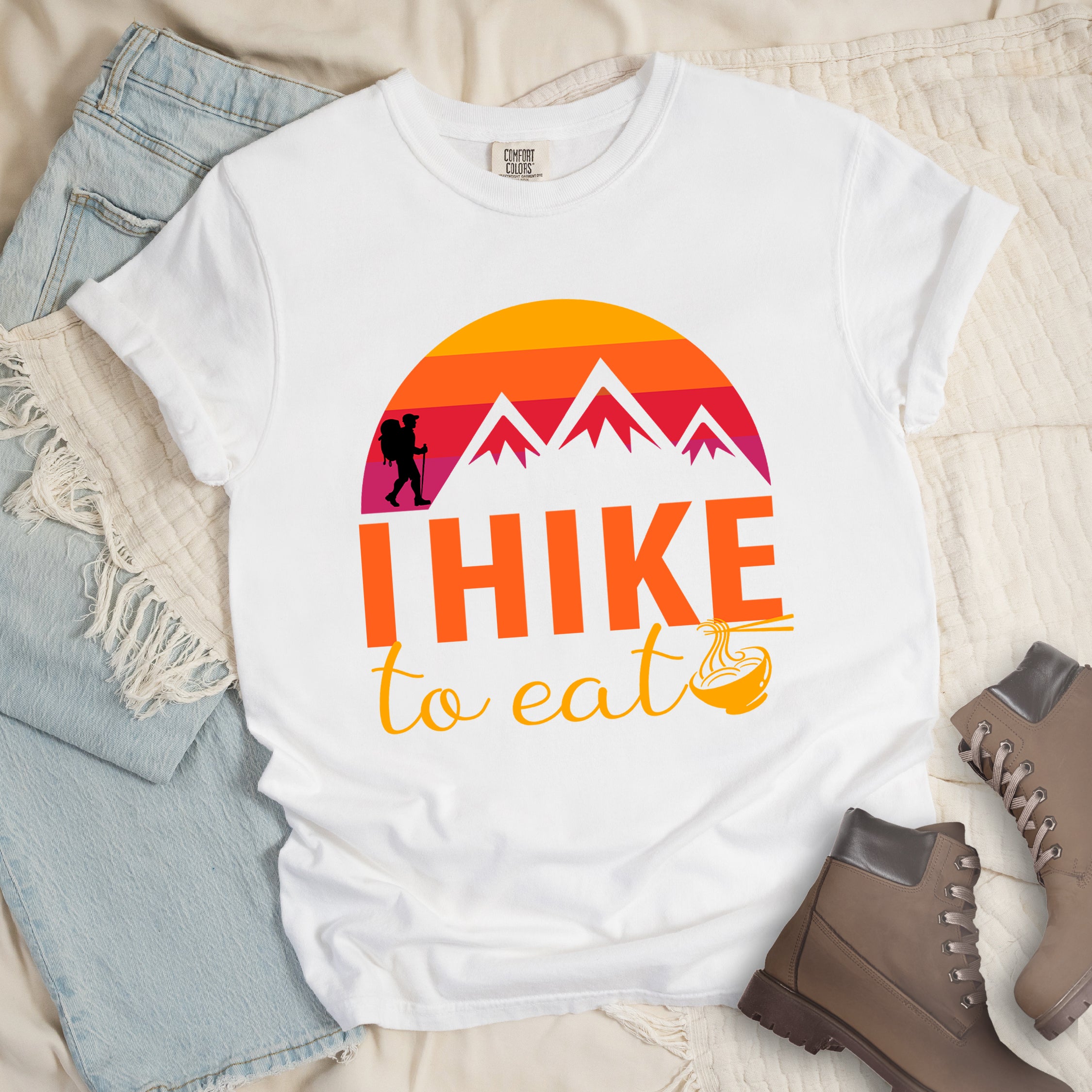White colored t-shirt featuring a retro sunset design with gradient colors from yellow to orange to red to purple. Mountain silhouettes and a small hiker figure appear against the sunset. Below this is "I HIKE" in large orange letters and "to eat" in yellow script, followed by a small illustration of a noodle bowl with chopsticks. The design has a vintage outdoor aesthetic. Features a classic crew neck and casual rolled sleeves, styled alongside light wash denim and brown hiking boots.