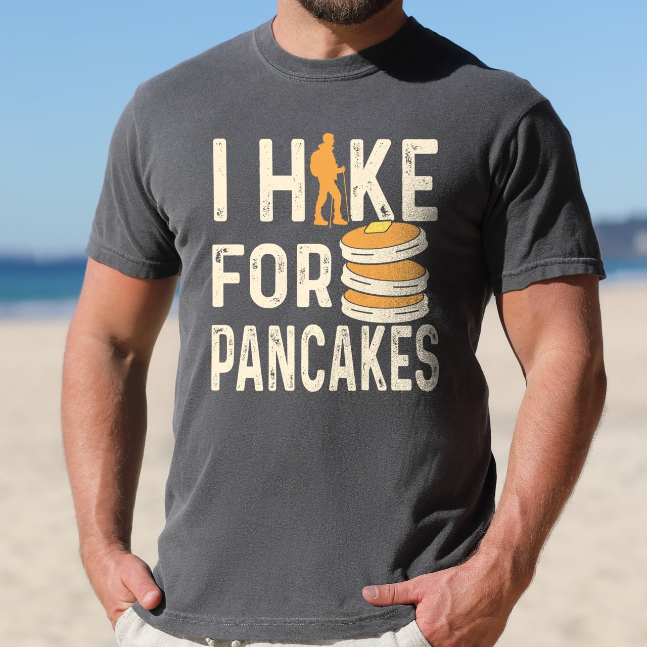 Man wearing Pepper (faded black) Pancake breakfast lover vintage tee I Hike for Pancakes with distressed cream typography character silhouette with trekking pole and illustrated stacked pancakes with butter on Comfort Colors shirt for brunch and food enthusiasts, hikers and foodies.