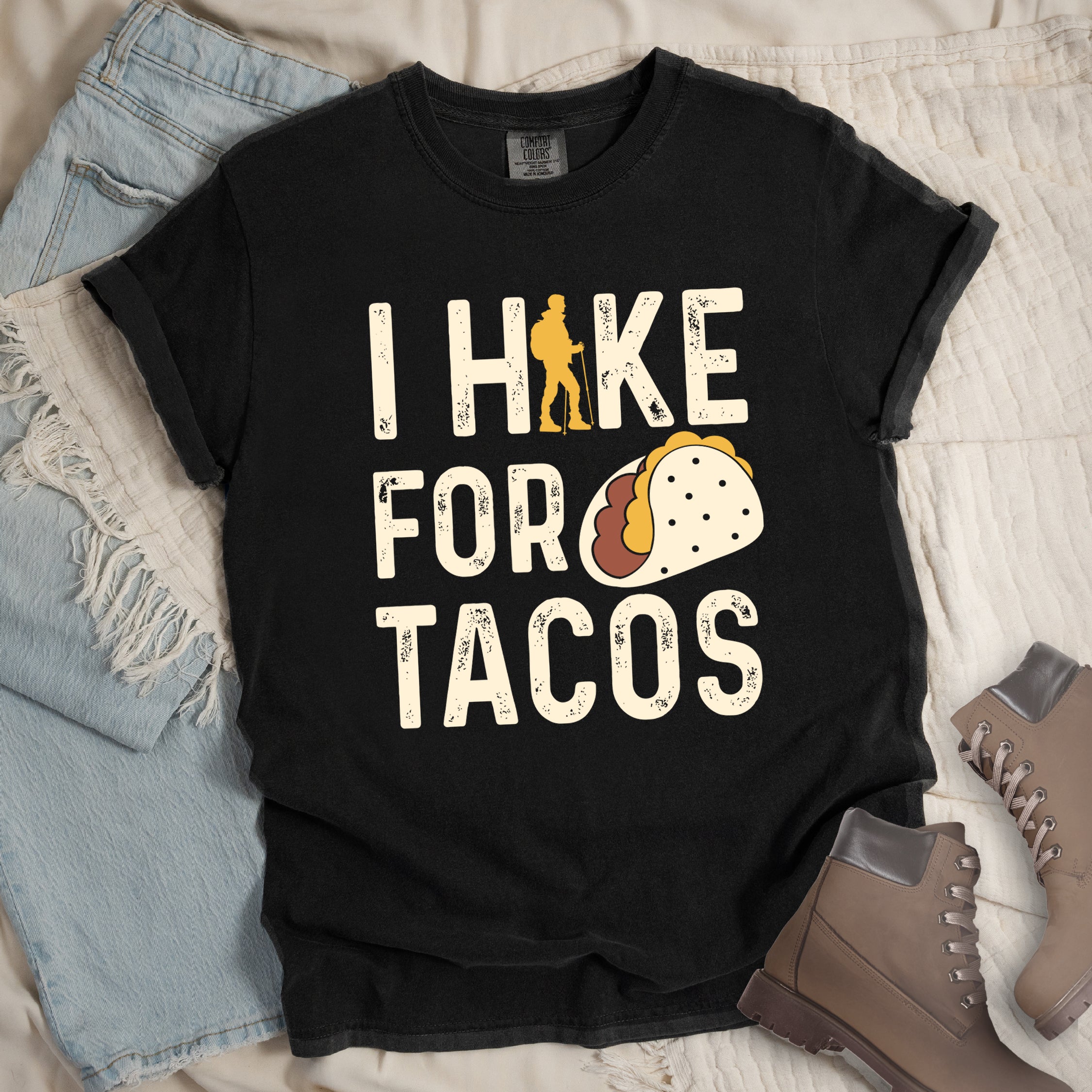 Black graphic t-shirt with 'I Hike for Tacos' text in cream lettering featuring orange hiker silhouette and colorful taco illustration, styled with light wash jeans and brown hiking boots.