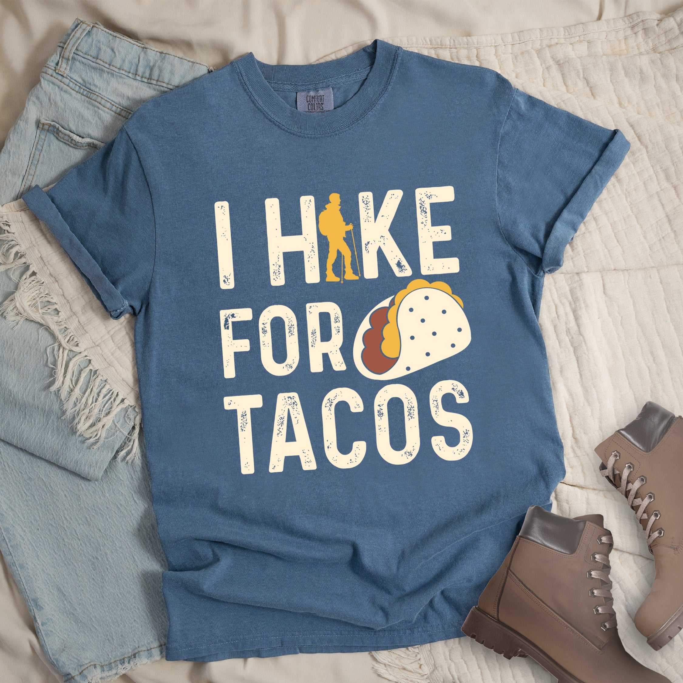 Blue Jean (Denim Blue) graphic t-shirt with 'I Hike for Tacos' text in cream lettering featuring orange hiker silhouette and colorful taco illustration, styled with light wash jeans and brown hiking boots.