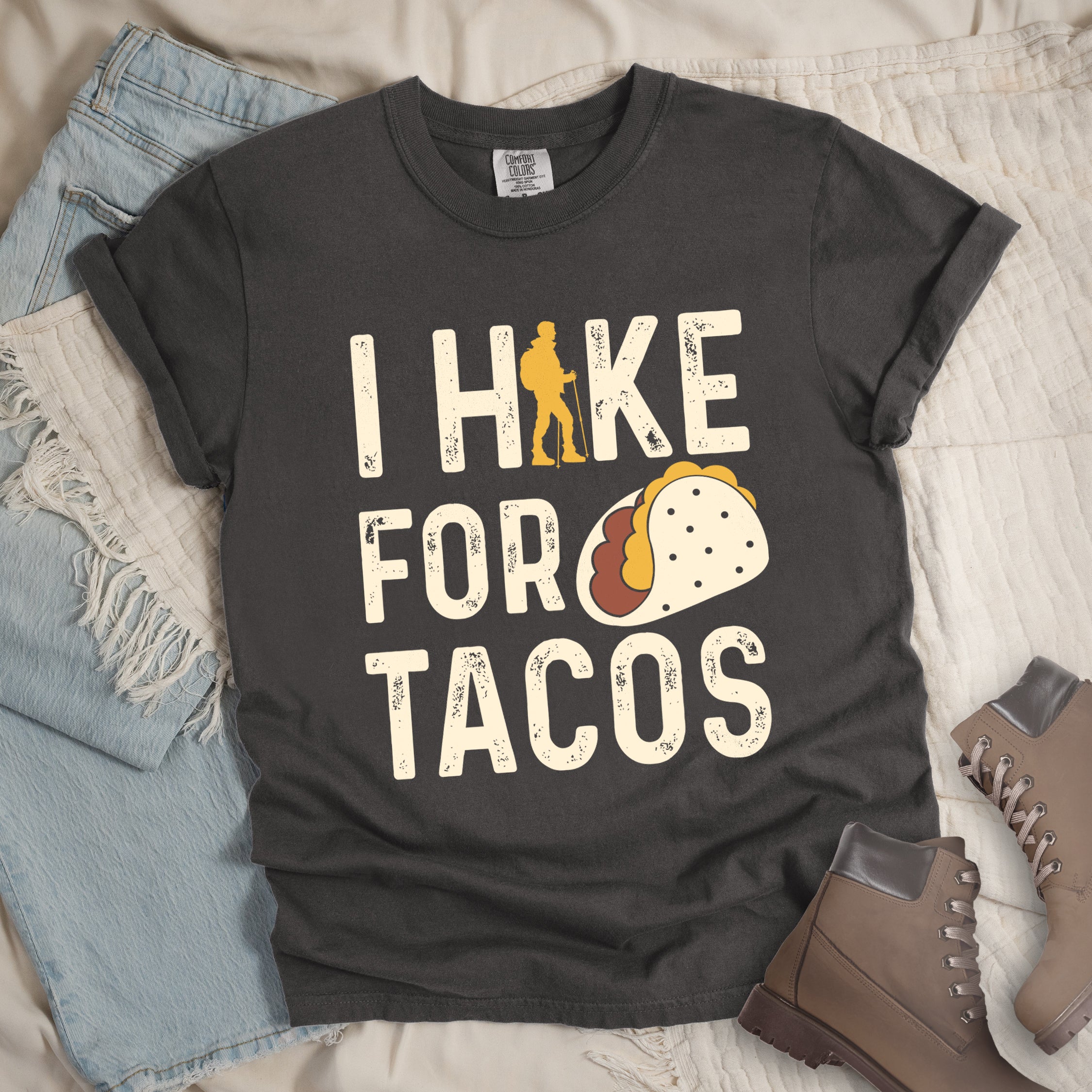 Pepper (Dark Gray) graphic t-shirt with 'I Hike for Tacos' text in cream lettering featuring orange hiker silhouette and colorful taco illustration, styled with light wash jeans and brown hiking boots