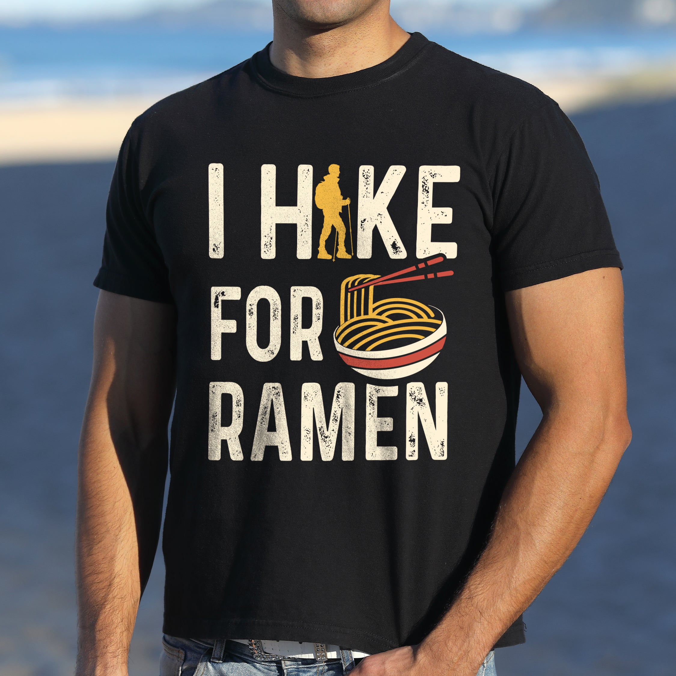 Black graphic t-shirt with 'I Hike for Ramen' text in cream lettering featuring orange hiker silhouette and colorful ramen bowl illustration with noodles and chopsticks.