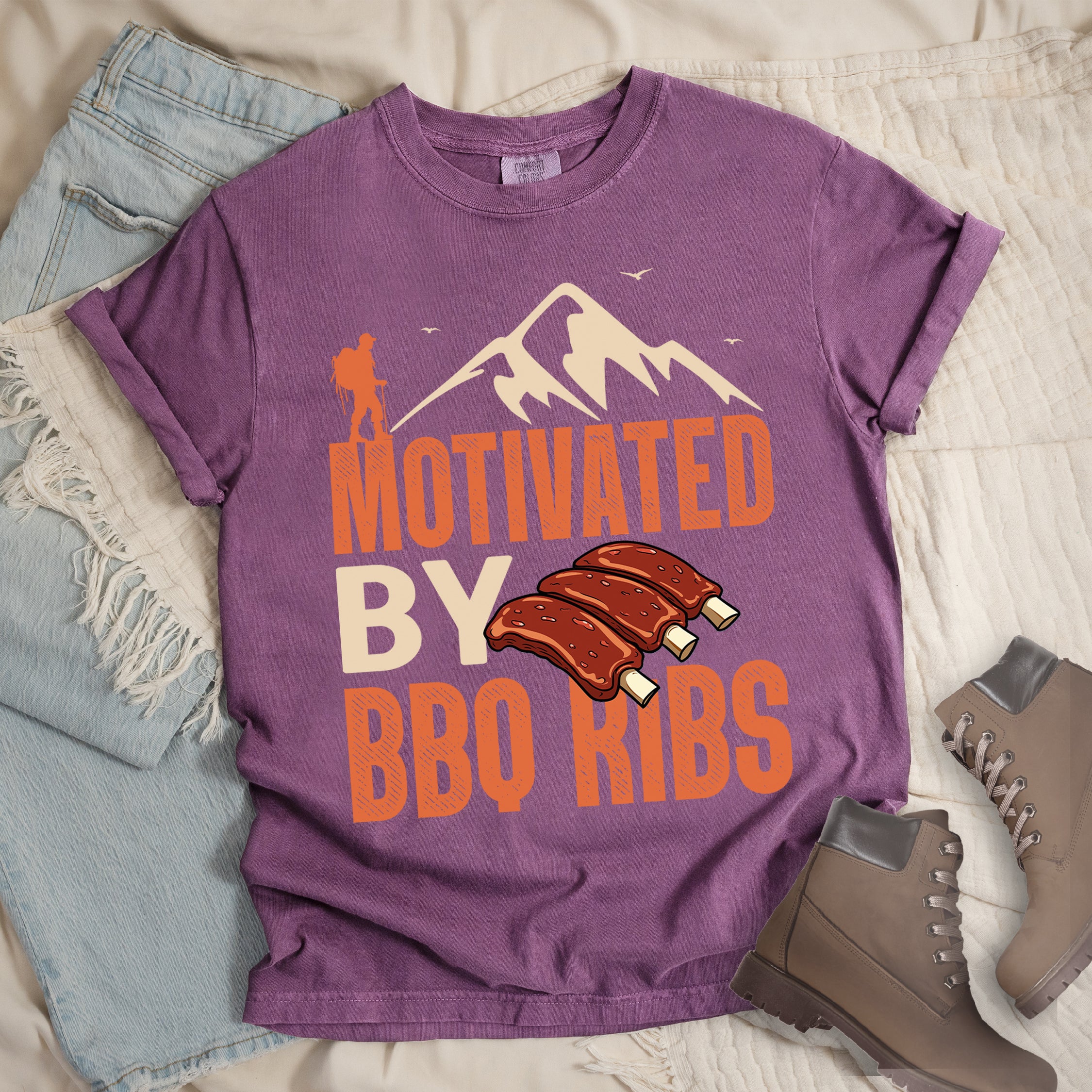 Berry (Dark Violet) t-shirt with text "MOTIVATED BY BBQ RIBS". At the top is a cream-colored mountain silhouette with flying birds and an orange hiker figure. "MOTIVATED" appears in large orange textured letters, "BY" in cream/tan, and "BBQ RIBS" in matching orange. Next to "BY" and above "BBQ RIBS" is an illustration of three barbecue ribs with exposed bones. The design combines hiking and food themes.