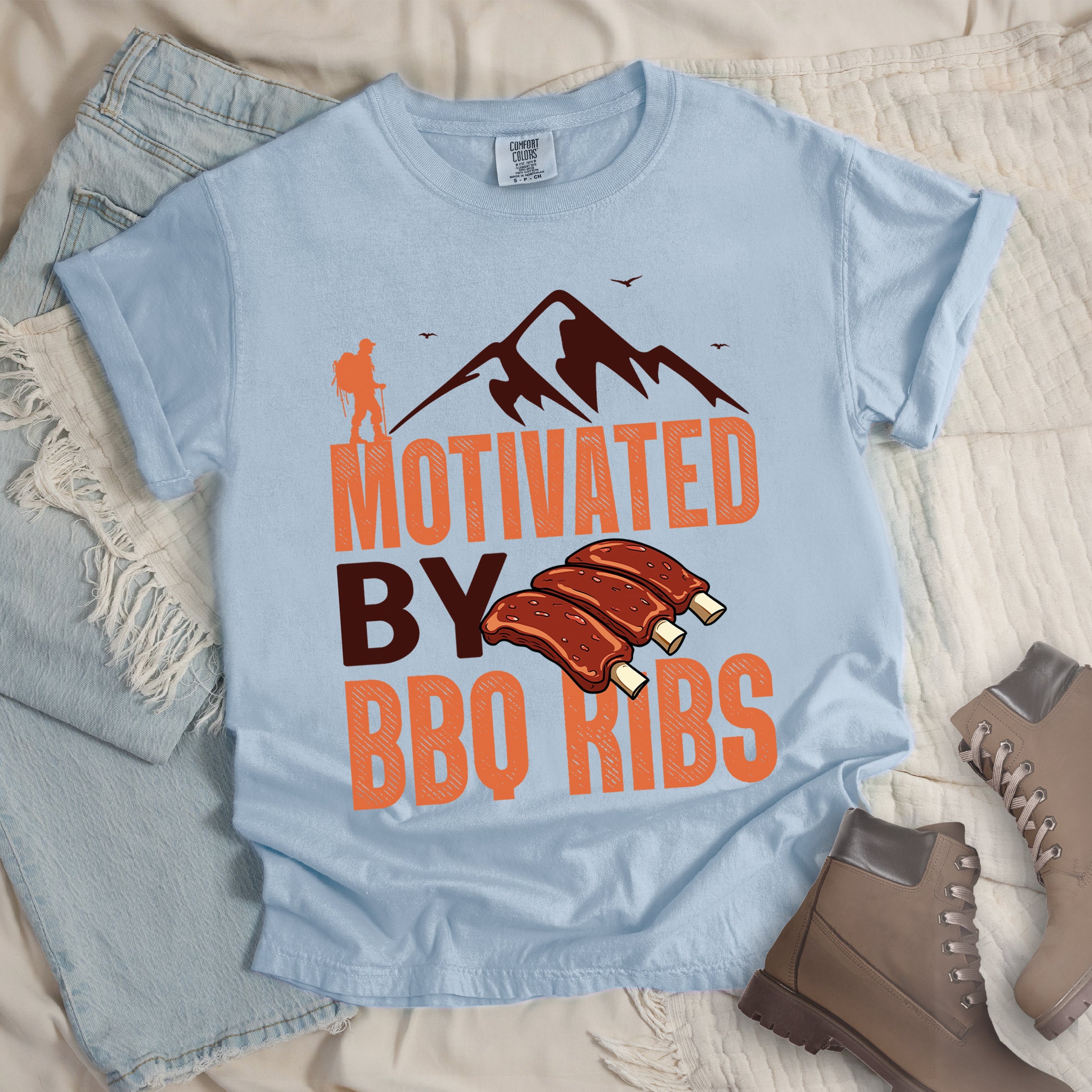 Chambray (Light Blue) t-shirt with text "MOTIVATED BY BBQ RIBS". At the top is a deep brown-colored mountain silhouette with flying birds and an orange hiker figure. "MOTIVATED" appears in large orange textured letters, "BY" in deep brown, and "BBQ RIBS" in matching orange. Next to "BY" and above "BBQ RIBS" is an illustration of three barbecue ribs with exposed bones. The design combines hiking and food themes.