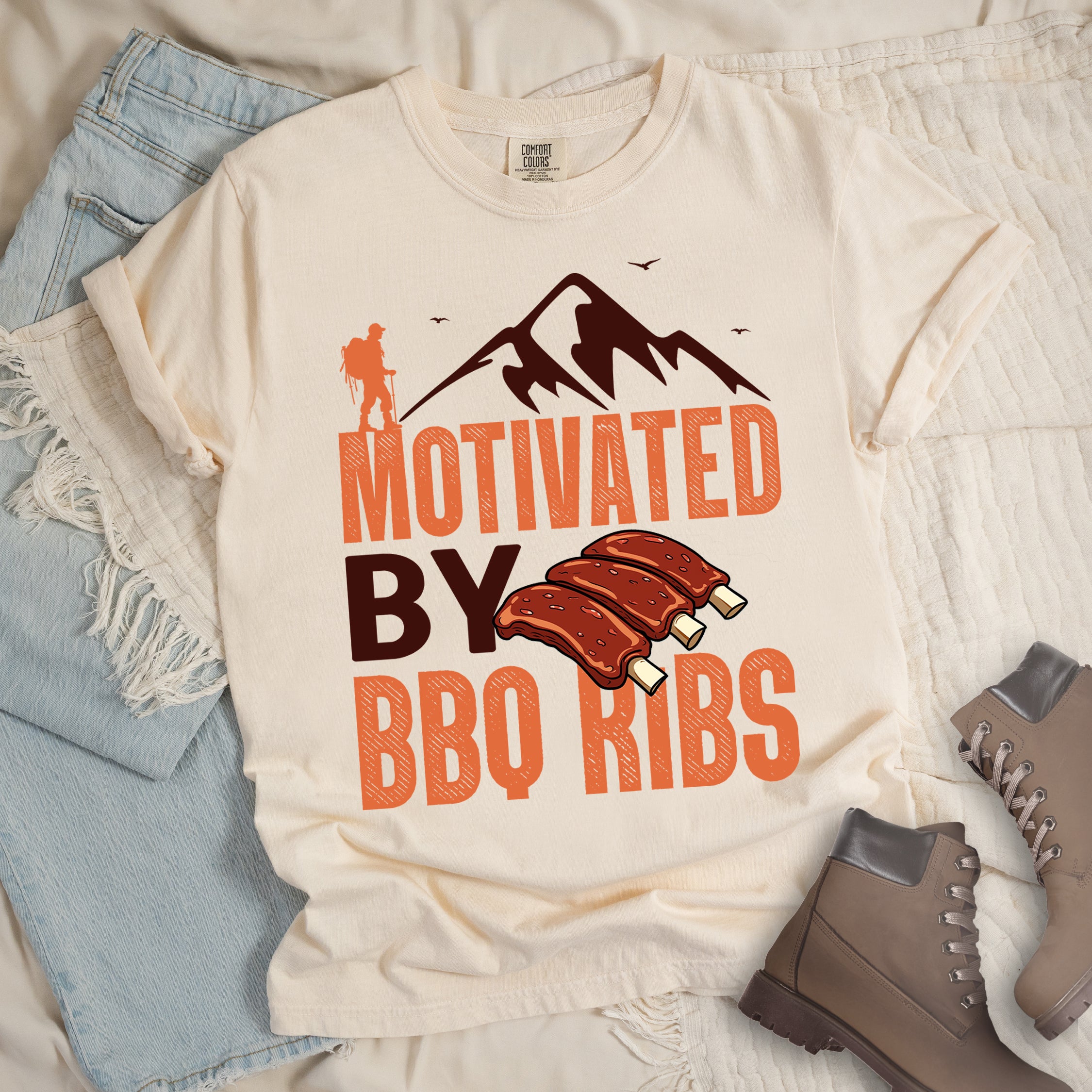 Ivory (Cream White) t-shirt with text "MOTIVATED BY BBQ RIBS". At the top is a deep brown-colored mountain silhouette with flying birds and an orange hiker figure. "MOTIVATED" appears in large orange textured letters, "BY" in deep brown, and "BBQ RIBS" in matching orange. Next to "BY" and above "BBQ RIBS" is an illustration of three barbecue ribs with exposed bones. The design combines hiking and food themes.