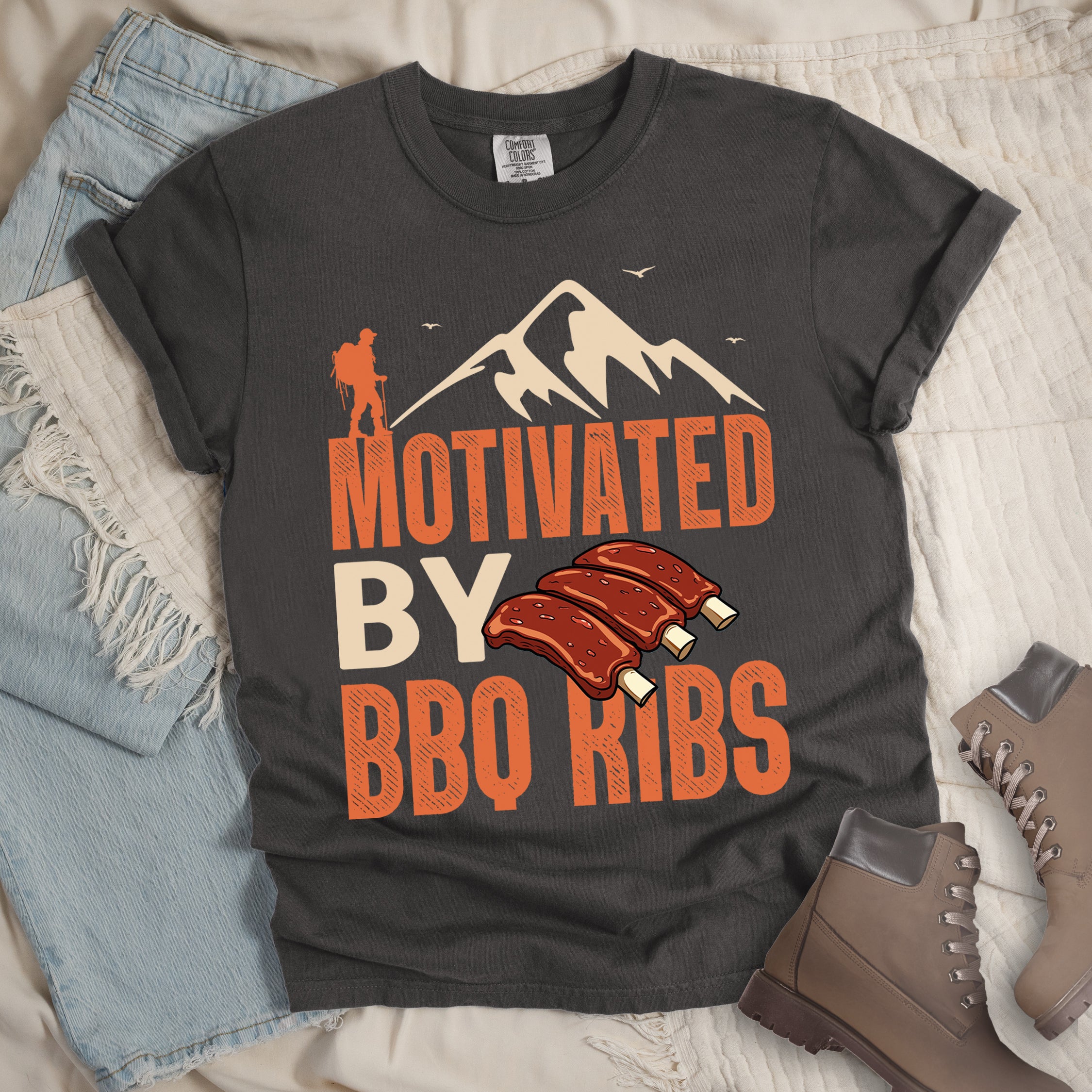 Pepper (Dark Gray) t-shirt with text "MOTIVATED BY BBQ RIBS". At the top is a cream-colored mountain silhouette with flying birds and an orange hiker figure. "MOTIVATED" appears in large orange textured letters, "BY" in cream/tan, and "BBQ RIBS" in matching orange. Next to "BY" and above "BBQ RIBS" is an illustration of three barbecue ribs with exposed bones. The design combines hiking and food themes.