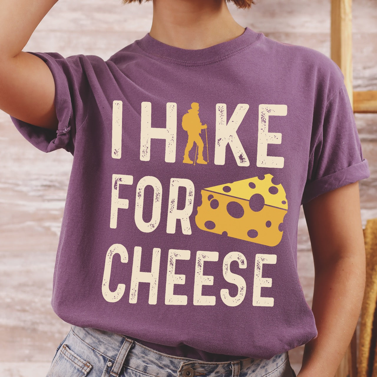 Woman wearing Purple Cheese lover vintage tee I Hike for Cheese with distressed cream typography character silhouette with trekking pole and illustrated Swiss cheese wedge with holes on Comfort Colors shirt for dairy and food enthusiasts, hikers and foodies.