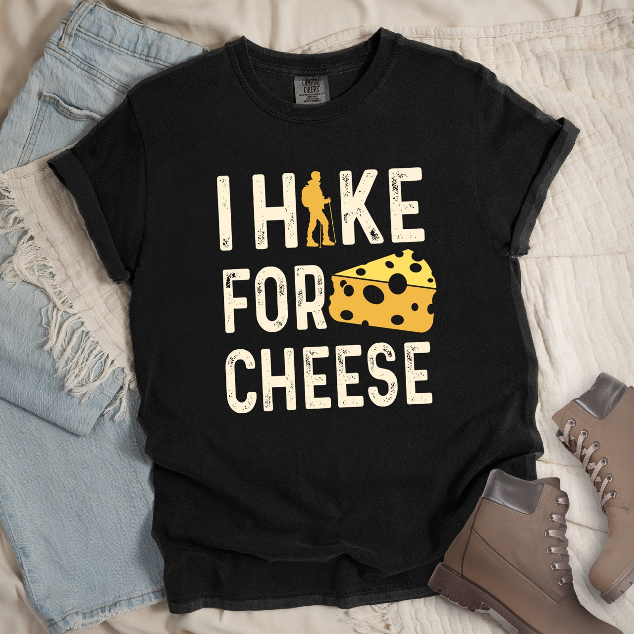 Black Cheese lover vintage tee I Hike for Cheese with distressed cream typography character silhouette with trekking pole and illustrated Swiss cheese wedge with holes on Comfort Colors shirt for dairy and food enthusiasts, hikers and foodies - laid flat with pants and boots