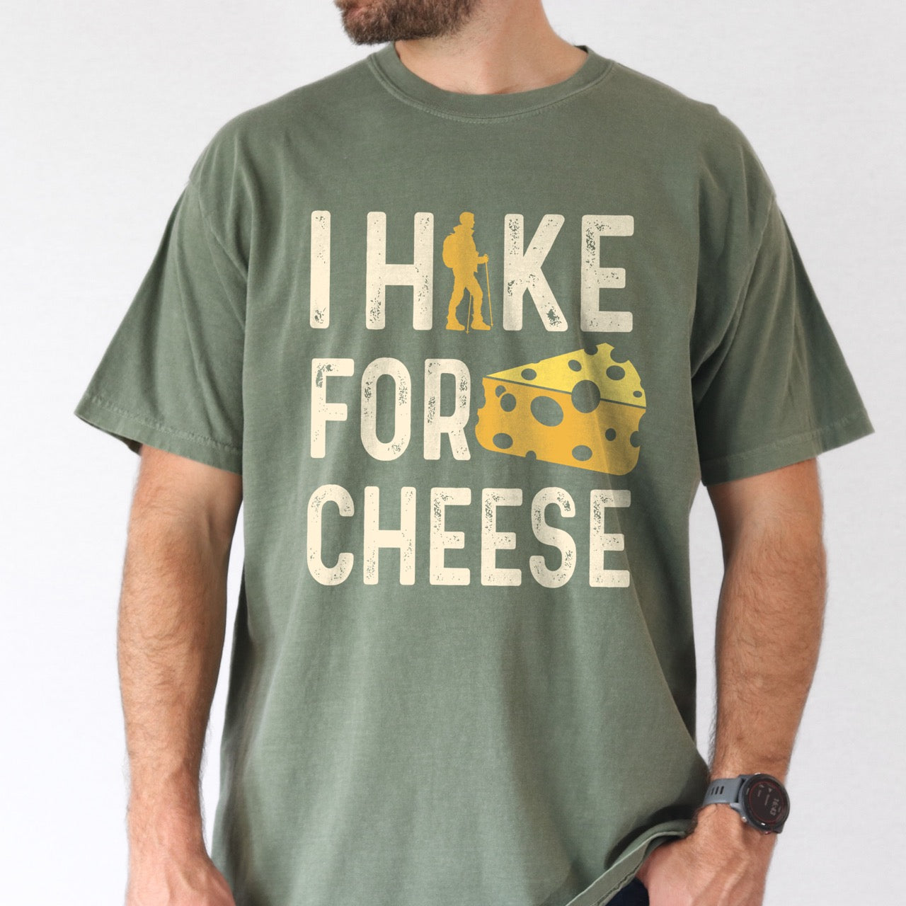 Man wearing Moss (dark green) Cheese lover vintage tee I Hike for Cheese with distressed cream typography character silhouette with trekking pole and illustrated Swiss cheese wedge with holes on Comfort Colors shirt for dairy and food enthusiasts, hikers and foodies.